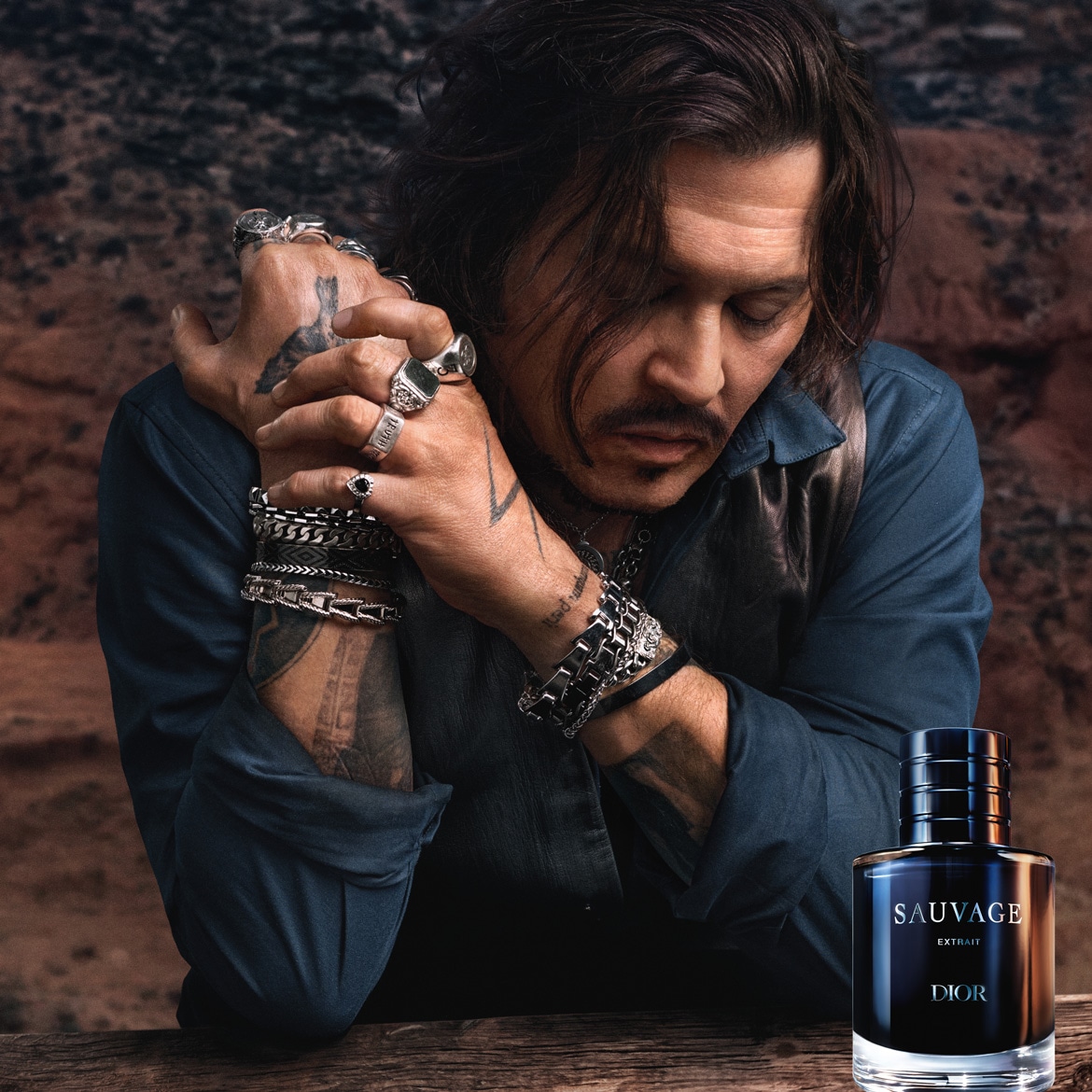 Johnny Depp, les yeux baissés, les mains jointes et ornées de bagues et de bracelets, pose à côté du parfum Dior Sauvage Extrait, un flacon en verre transparent avec un liquide bleu foncé et un capuchon noir, sur une table en bois.