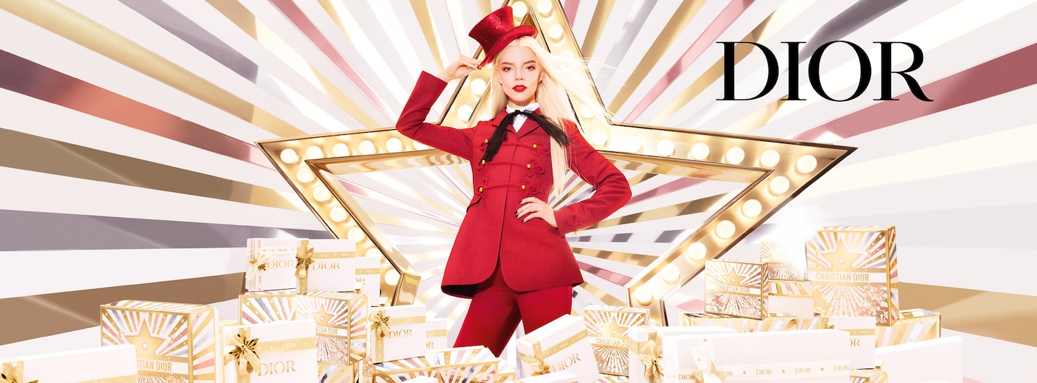 Une femme en costume rouge et chapeau haut de forme pose devant une étoile lumineuse, entourée de cadeaux Dior aux motifs éclatants.'