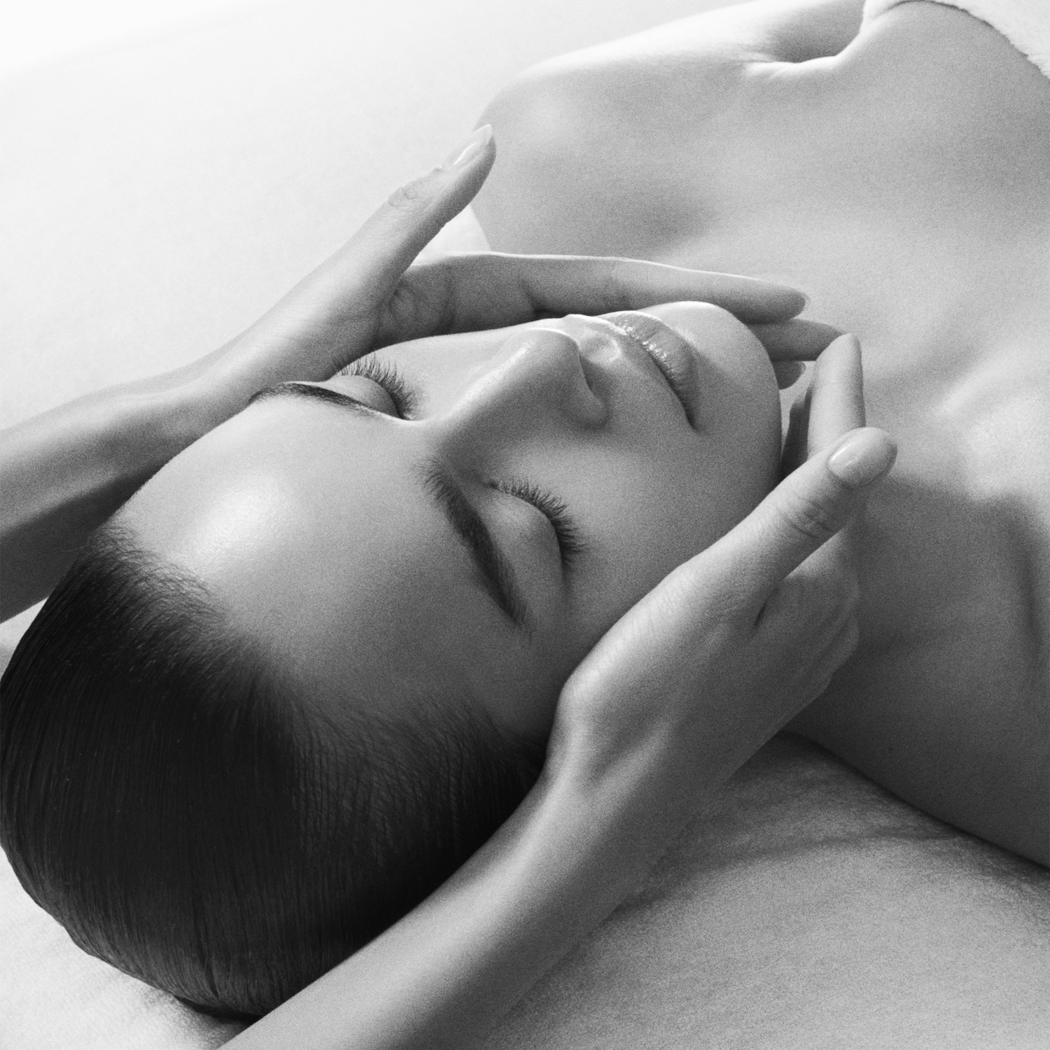 Gros plan en noir et blanc d'une femme allongée sur le dos, les yeux fermés, tandis que des mains massent doucement son visage, offrant une sensation de détente et de soin.