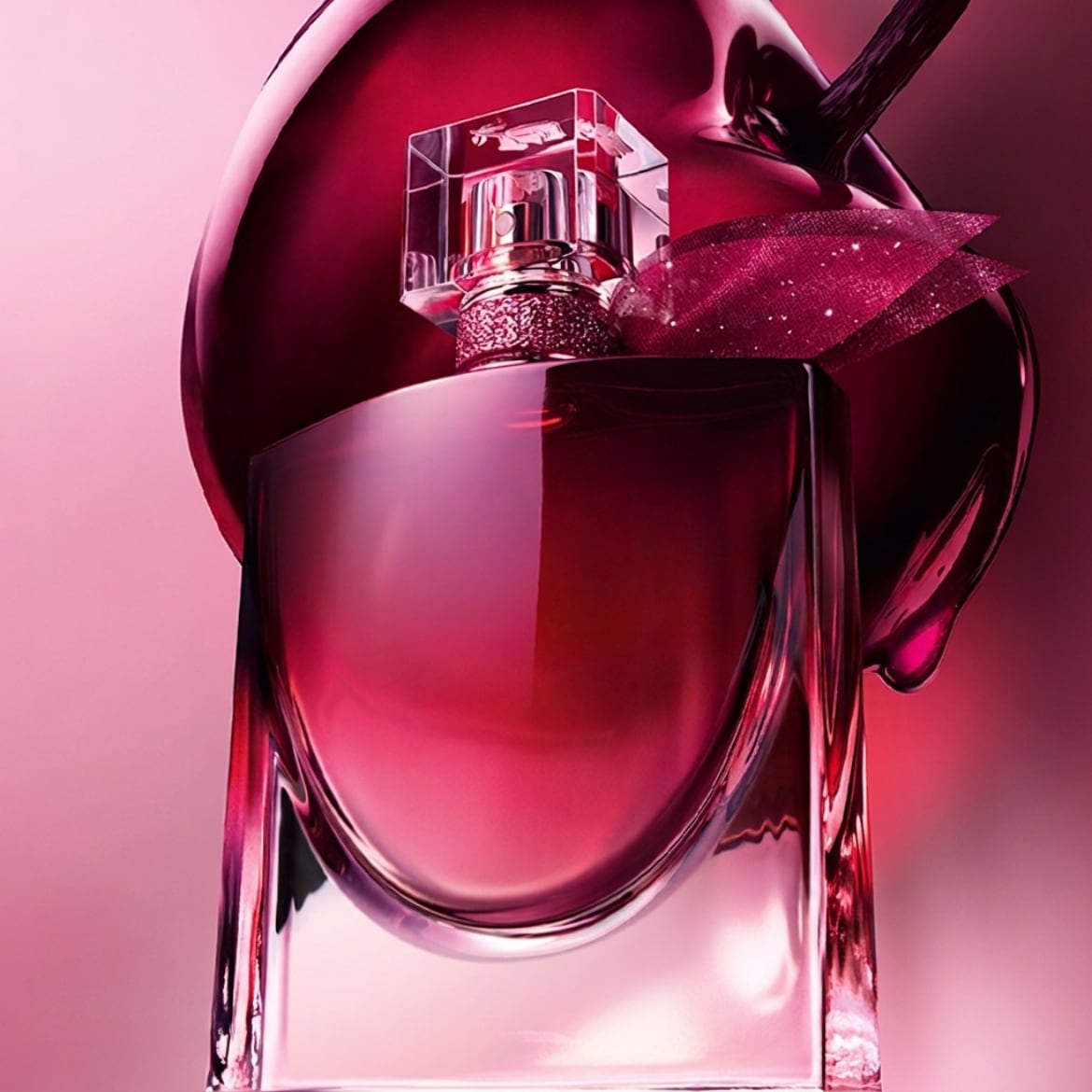 Le parfum La Vie Est Belle de Lancôme se présente dans un flacon en verre transparent avec un liquide rouge-rose, orné d'un ruban scintillant et d'un bouchon en verre, sur un fond rose vif.