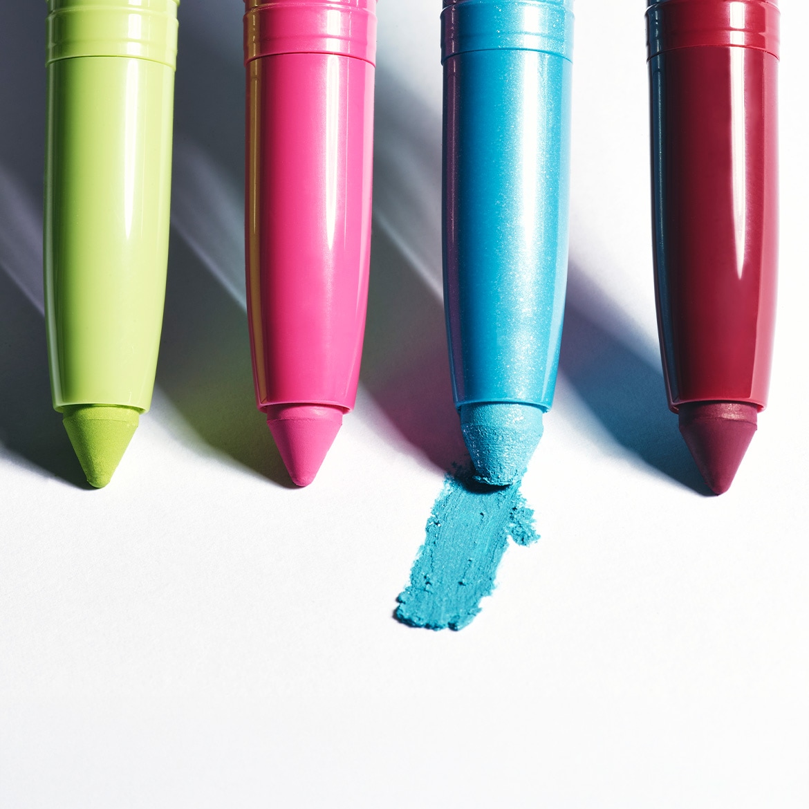 Quatre crayons de maquillage aux couleurs vives, vert citron, rose vif, bleu turquoise et bordeaux, sont disposés verticalement sur un fond blanc, le crayon bleu ayant laissé une trace de couleur.