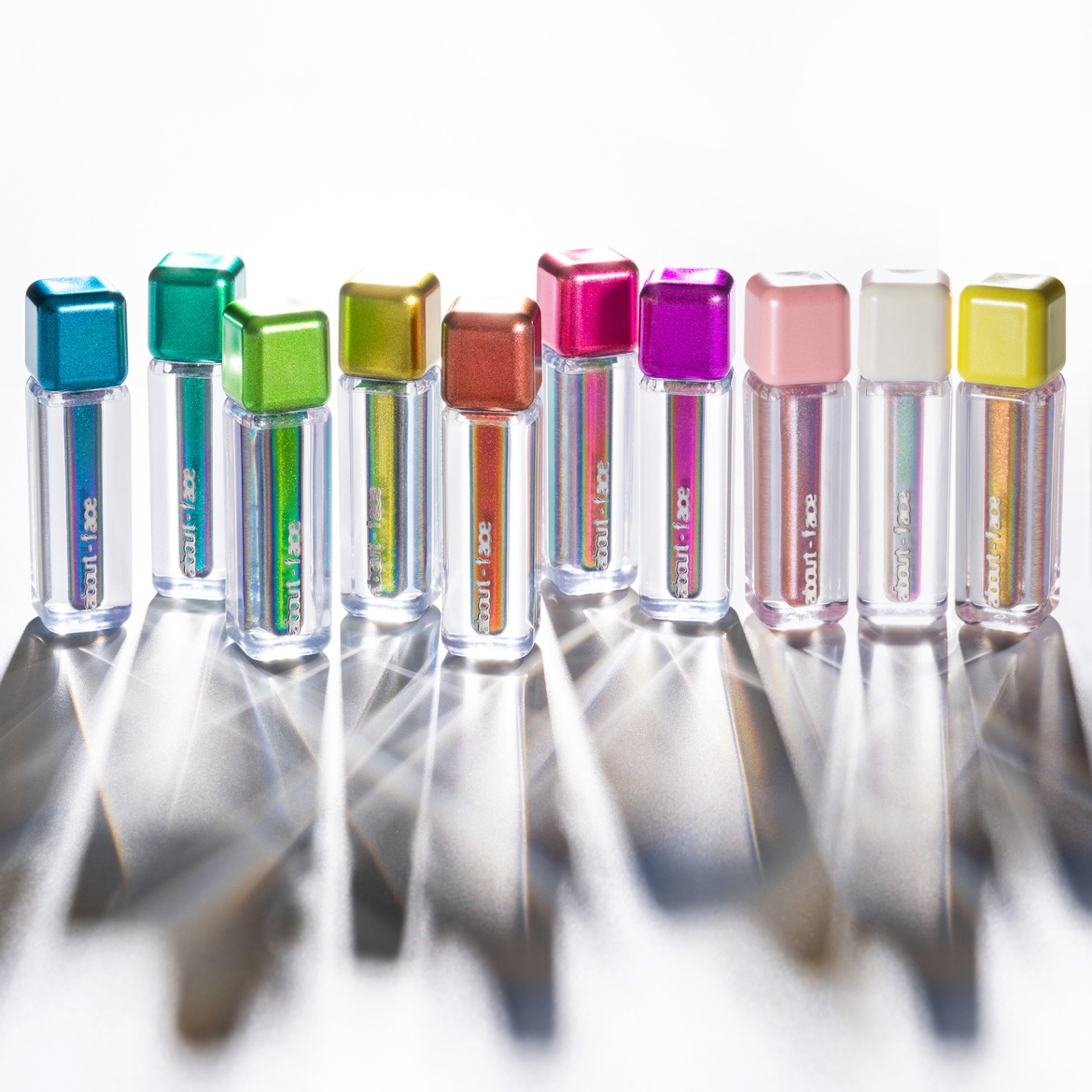 Découvrez la gamme de brillants à lèvres About-Face, présentés dans des flacons transparents avec des bouchons colorés allant du bleu au jaune, offrant une brillance éclatante pour chaque humeur.