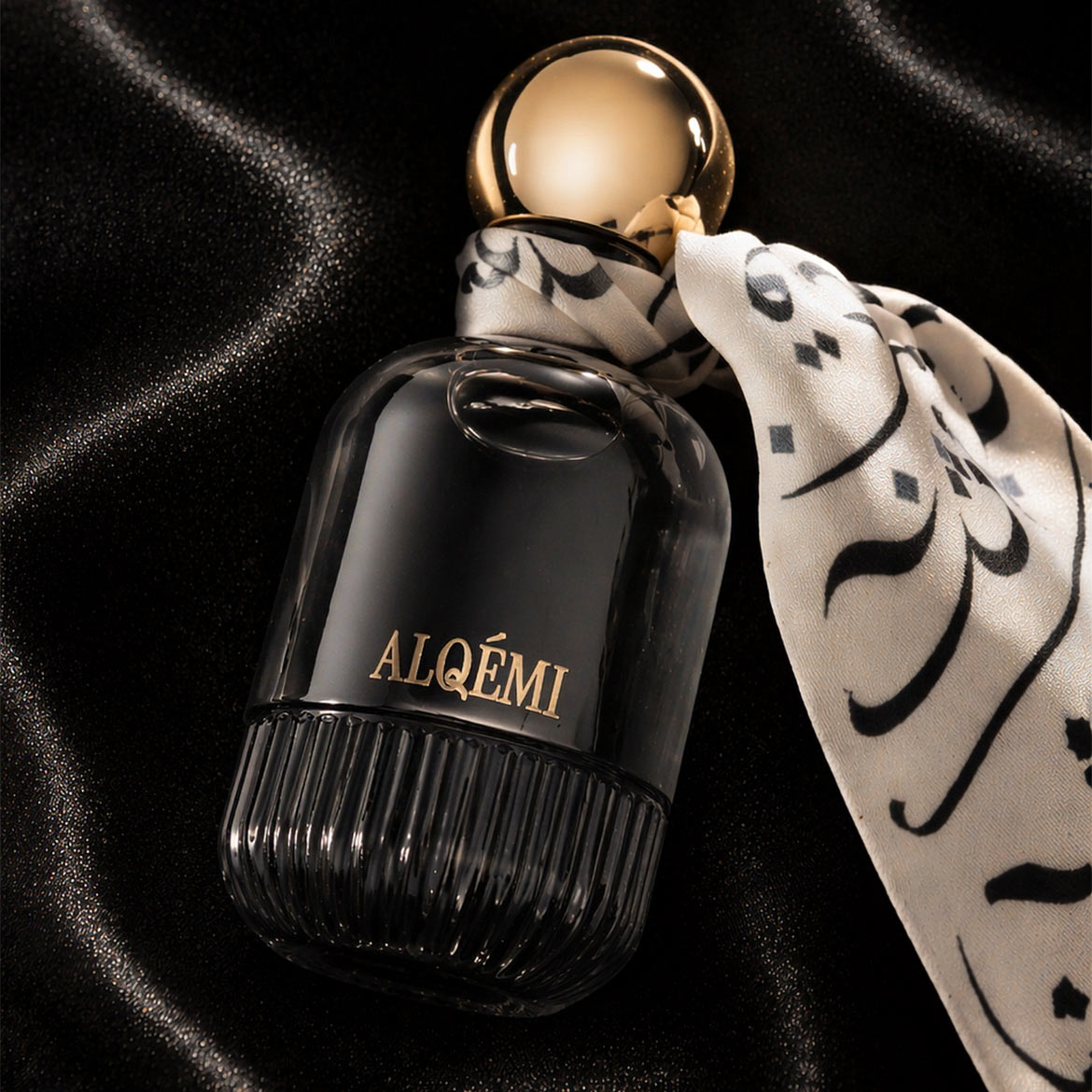Le parfum Alqémi est présenté dans un flacon en verre noir avec un bouchon doré, orné d'un ruban blanc et noir, posé sur un tissu noir soyeux.