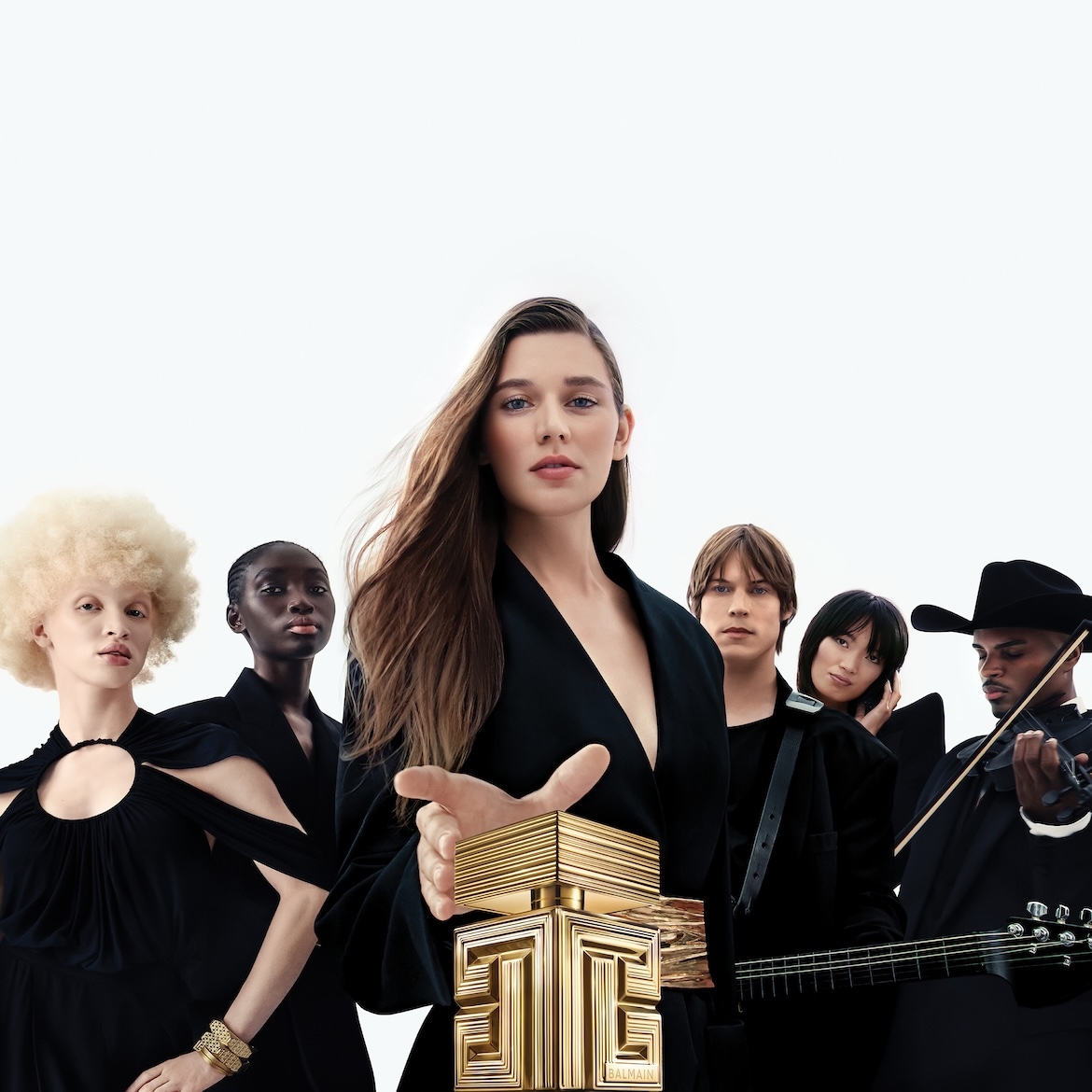 Une femme aux cheveux longs et bruns, vêtue d'une veste noire, tient un flacon de parfum doré Balmain, entourée de quatre autres personnes aux styles variés, sur un fond blanc.