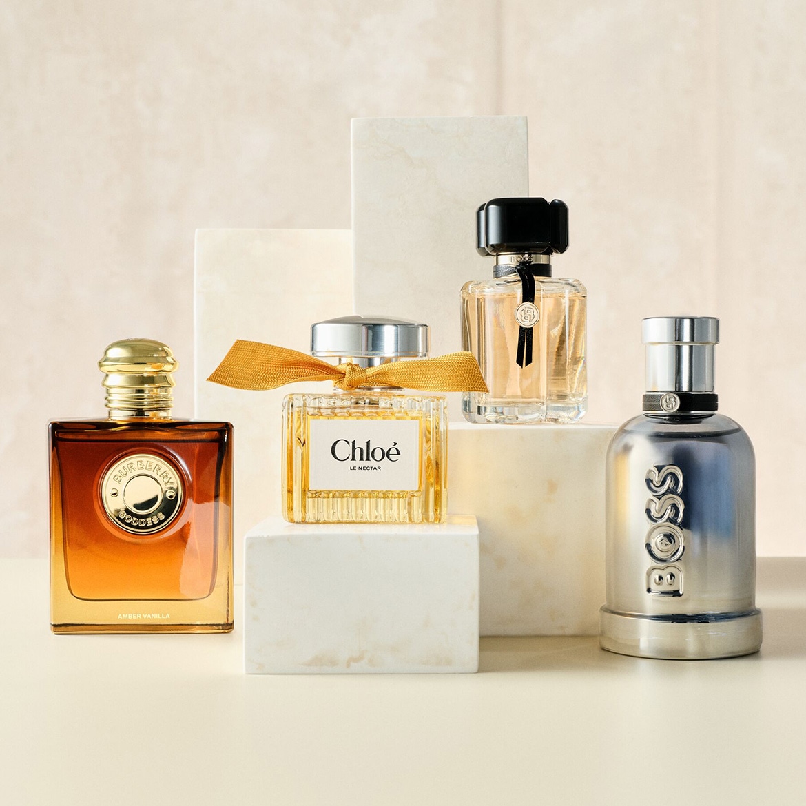 Quatre flacons de parfum de différentes marques, dont Burberry Goddess, Chloé Le Nectar et Boss, sont présentés sur des blocs de marbre blanc, avec des teintes ambrées et dorées.