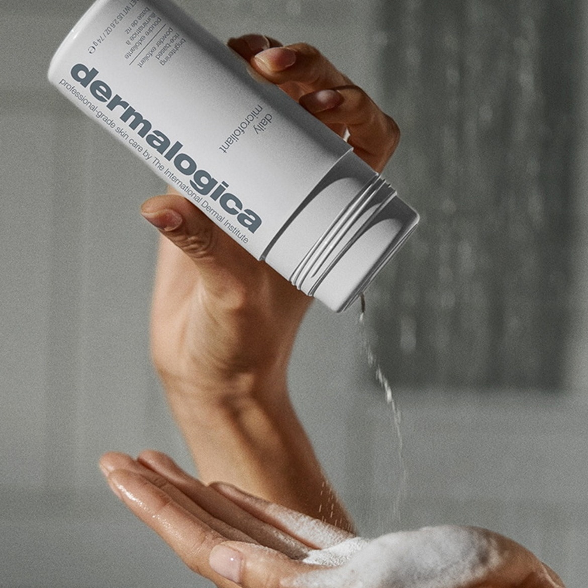 Une main tient une bouteille blanche de Dermalogica Daily Microfoliant, une poudre exfoliante éclaircissante à base de riz, et verse le produit dans l'autre main, créant une mousse blanche.