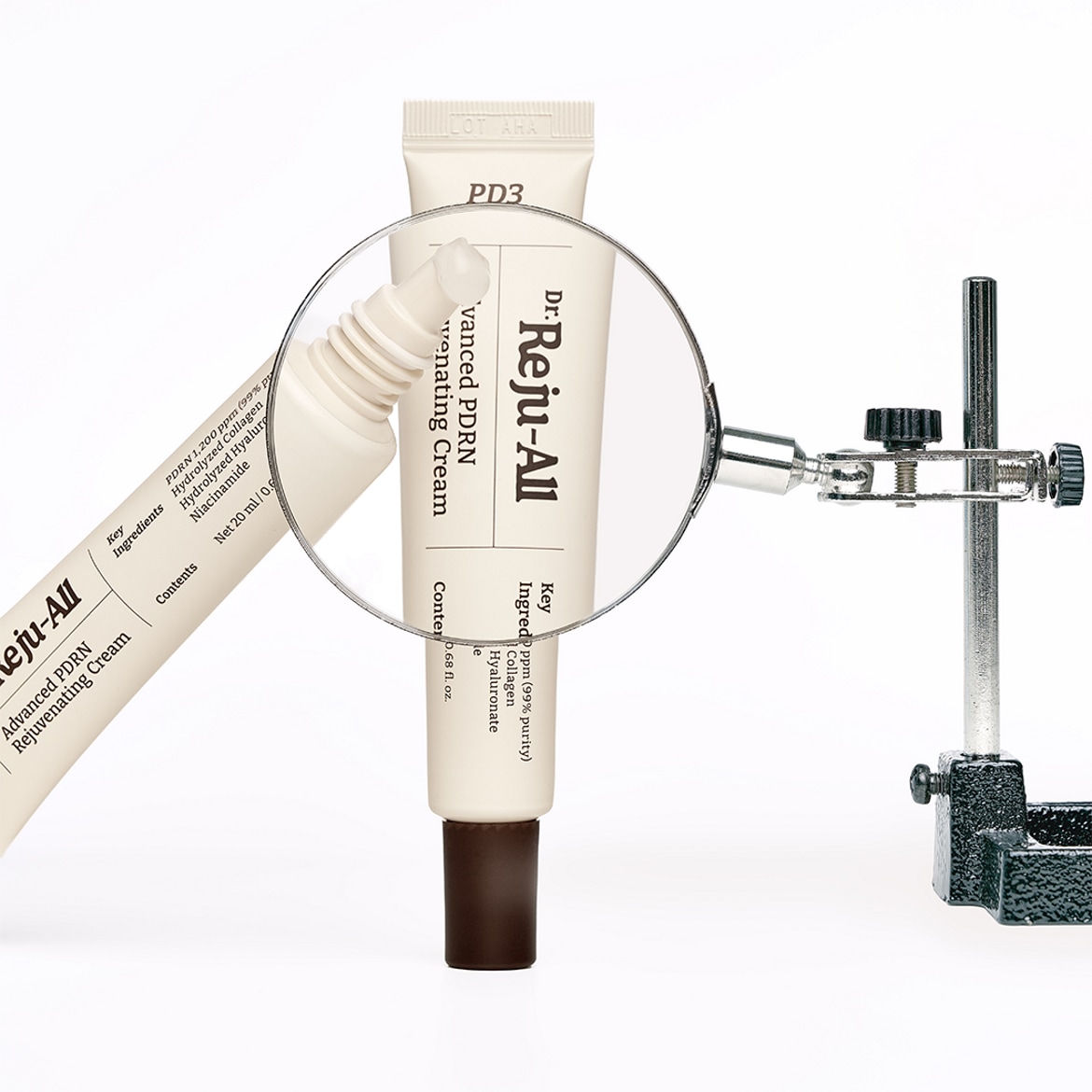 Le Dr. Reju-All Advanced PDRN Rejuvenating Cream, un tube blanc de 20 ml avec un bouchon marron, est mis en évidence par une loupe, révélant les ingrédients clés tels que le PDRN, le collagène hydrolysé, l'hyaluronate et la niacinamide.