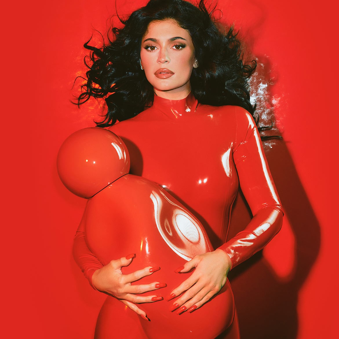 Kylie Jenner, vêtue d'une combinaison rouge brillante, pose avec une forme abstraite rouge, le tout sur un fond rouge uni.