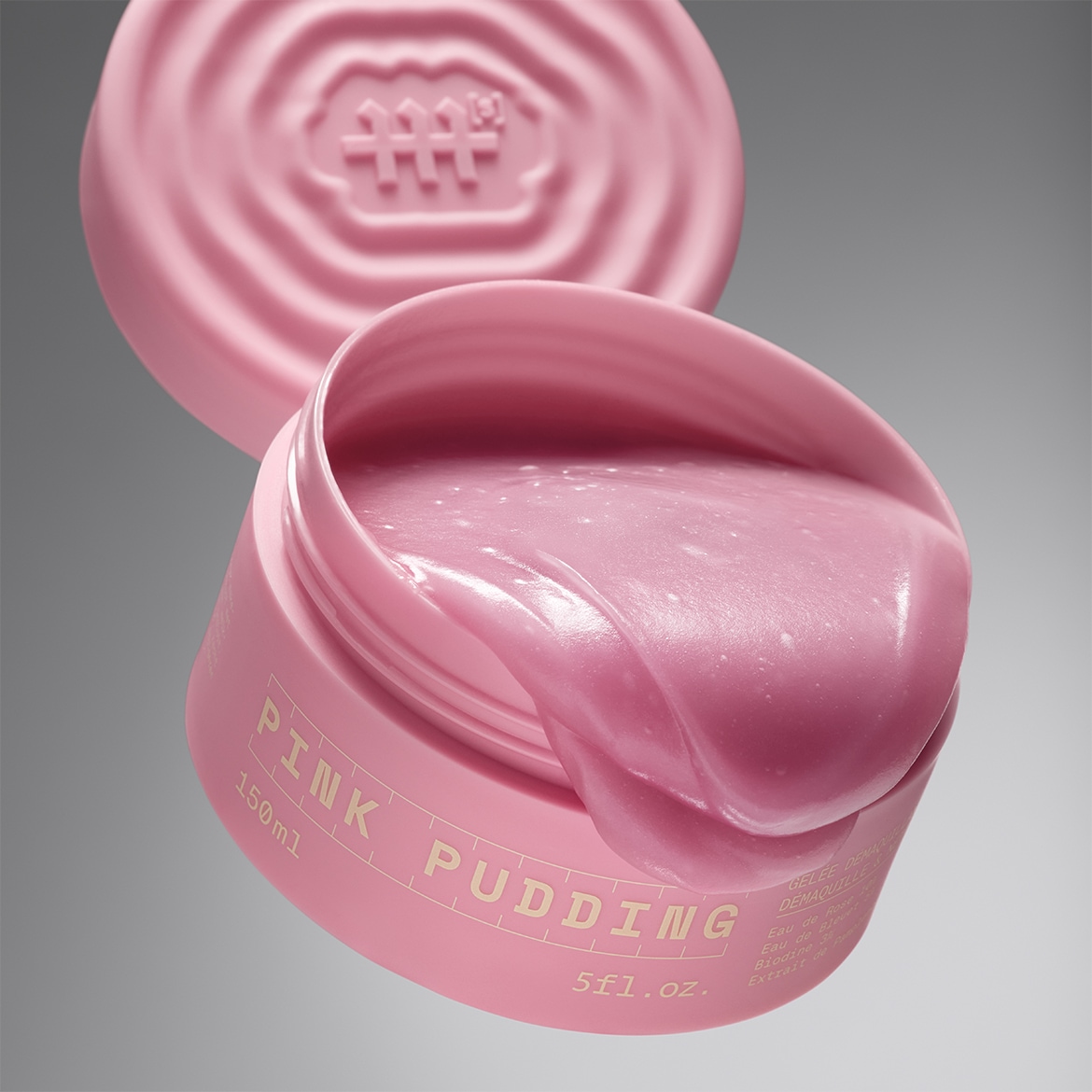 Le pot de 150 ml de Pink Pudding, un démaquillant rose vif à la texture gélatineuse, est ouvert, révélant son contenu riche et crémeux, avec le couvercle posé derrière.