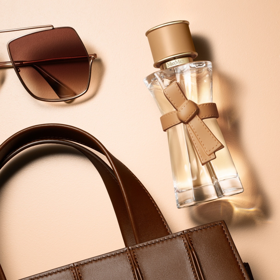 Un flacon de parfum Max Mara en verre transparent avec un ruban en cuir beige, posé à côté de lunettes de soleil marron et d'un sac en cuir marron, le tout sur un fond beige clair.