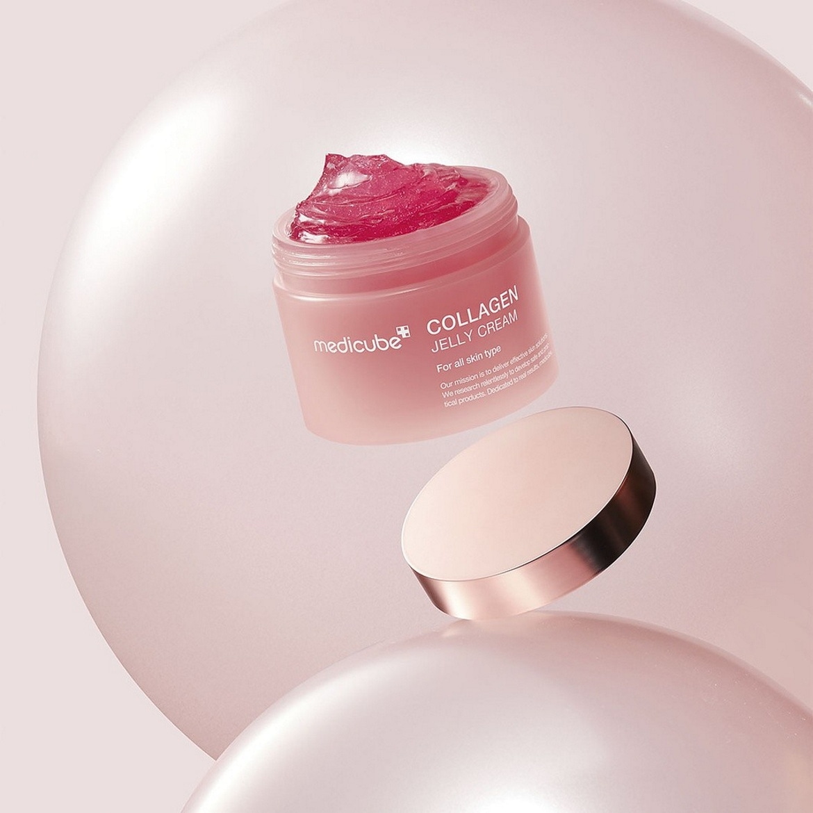 Le pot de crème Medicube Collagen Jelly, de couleur rose pâle, est ouvert et révèle une texture gélatineuse rose vif, avec son couvercle rose doré posé à côté, le tout sur un fond rose clair.