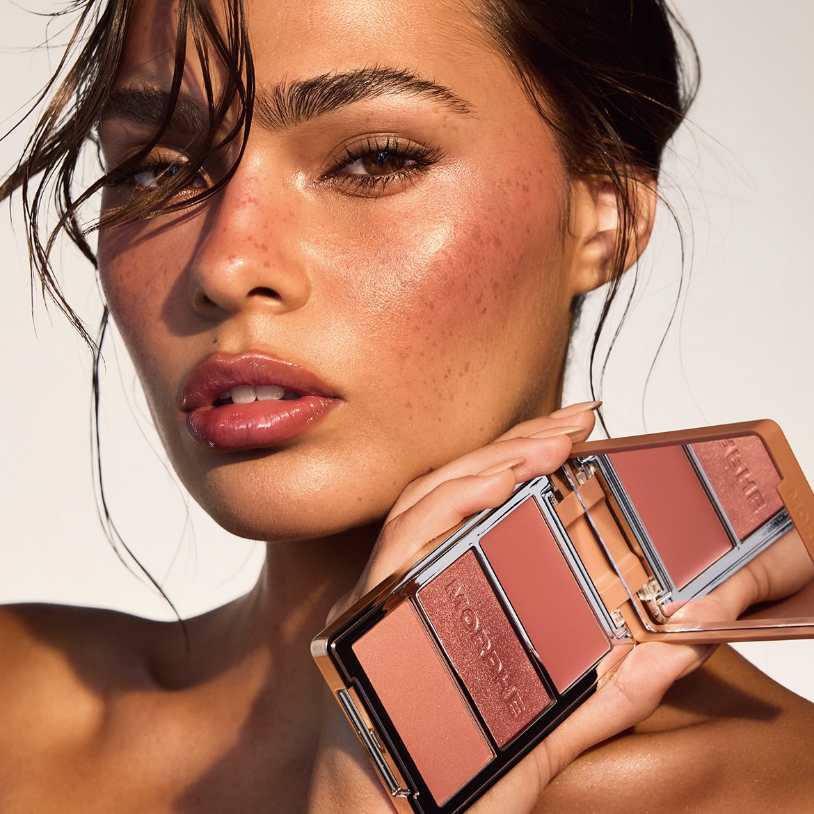 Une femme au teint éclatant et aux joues rosées tient une palette de maquillage Morphe avec des fards à joues et des illuminateurs dans des tons pêche et bronze.