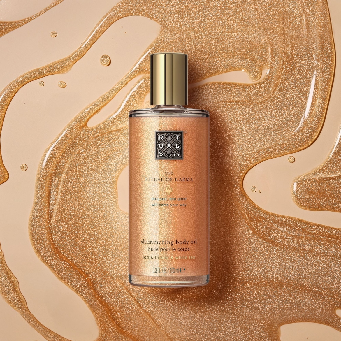 L'huile corporelle chatoyante The Ritual of Karma de 100 ml, infusée de fleur de lotus et de thé blanc, est présentée dans une bouteille en verre avec un bouchon doré, posée sur un fond beige avec des traînées d'huile dorée scintillante.