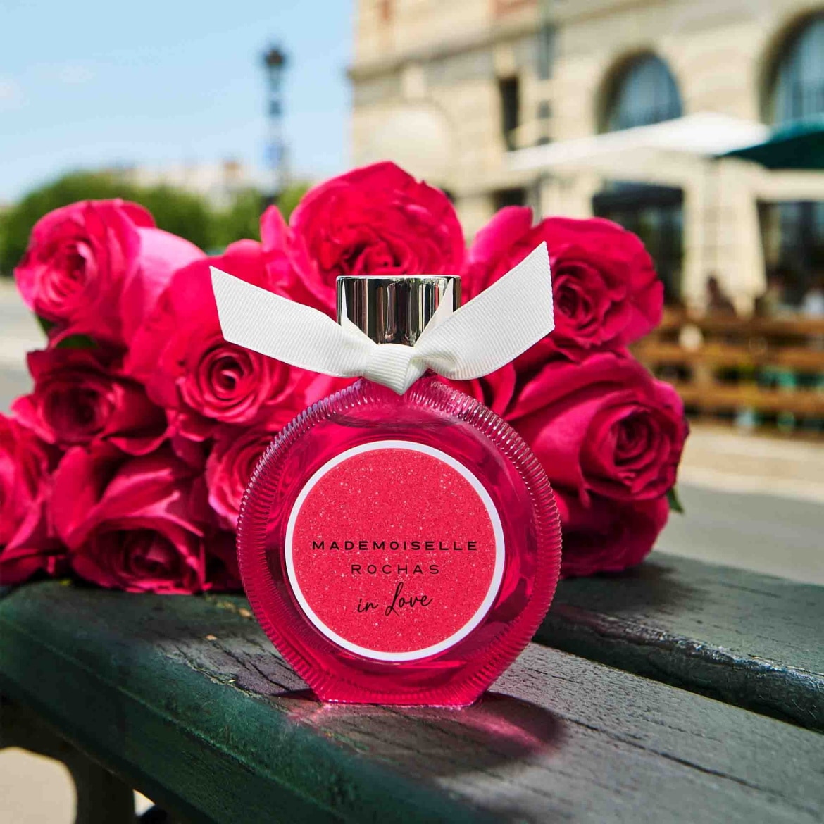 Découvrez Mademoiselle Rochas In Love, un parfum floral et fruité présenté dans un flacon rose orné d'un ruban blanc, sur un banc avec un bouquet de roses rouges.