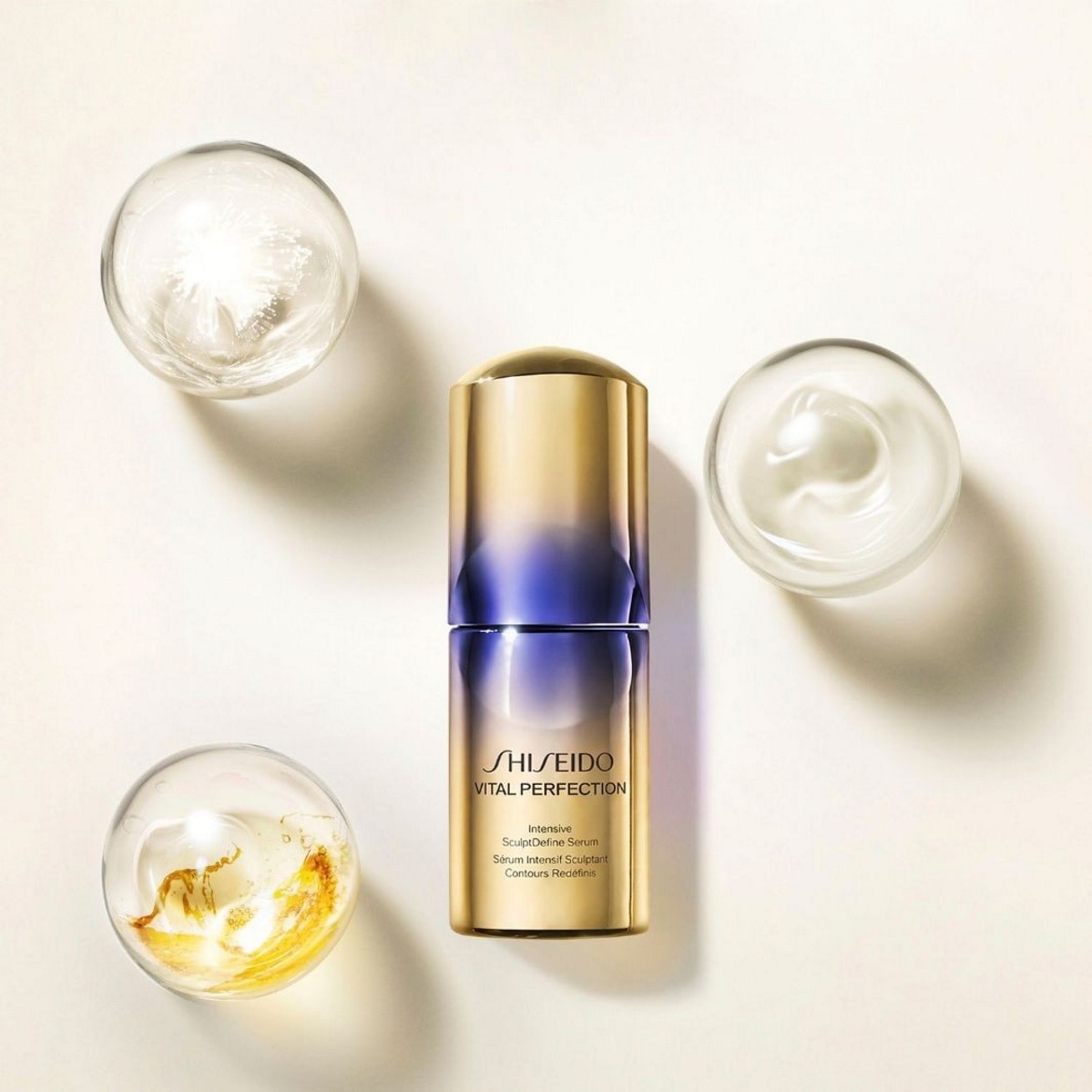 Le sérum Shiseido Vital Perfection Intensive SculptDefine, dans un flacon doré et violet, est présenté avec trois coupelles en verre contenant des textures de sérum variées, illustrant ses ingrédients et sa consistance.
