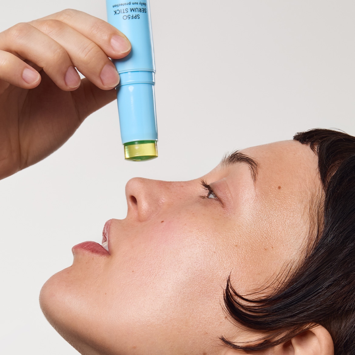 Une femme applique un stick solaire SPF50 sur son visage, le stick est bleu clair et laisse une texture brillante sur la peau.