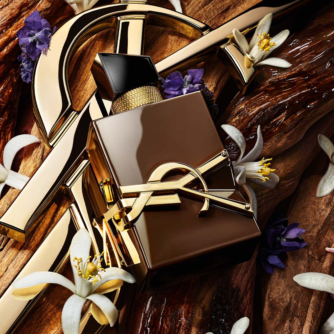 Un flacon de parfum marron foncé avec un logo doré YSL est posé sur un fond en bois texturé, orné de fleurs blanches et violettes.