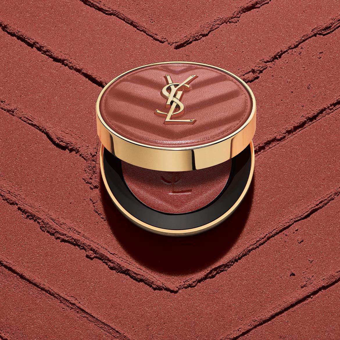 Un fard à paupières Yves Saint Laurent ouvert, de couleur marron-rouge, est posé sur une surface texturée de la même couleur, avec le logo YSL doré sur le couvercle.