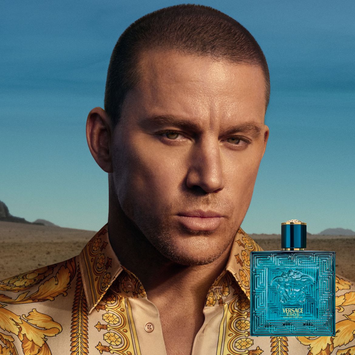 Channing Tatum présente le parfum Versace Eros, un flacon rectangulaire bleu turquoise avec un motif en relief et un bouchon assorti, sur fond de paysage désertique.