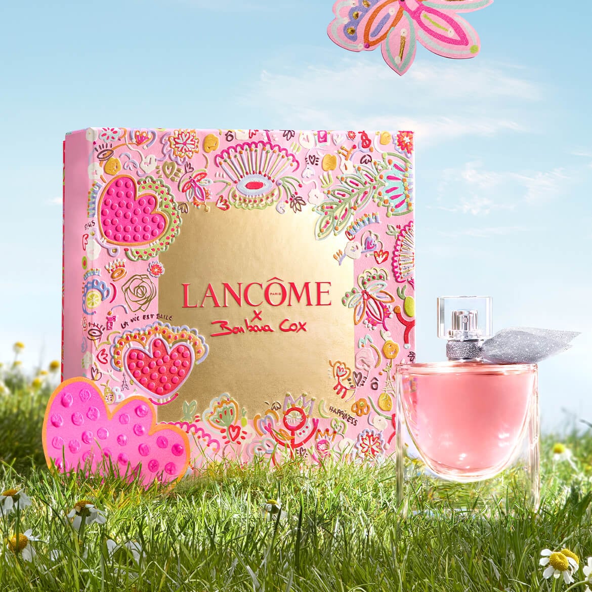 Découvrez le parfum Lancôme x Barbara Cox dans un coffret rose orné de motifs floraux et de cœurs, présenté sur une pelouse ensoleillée.
