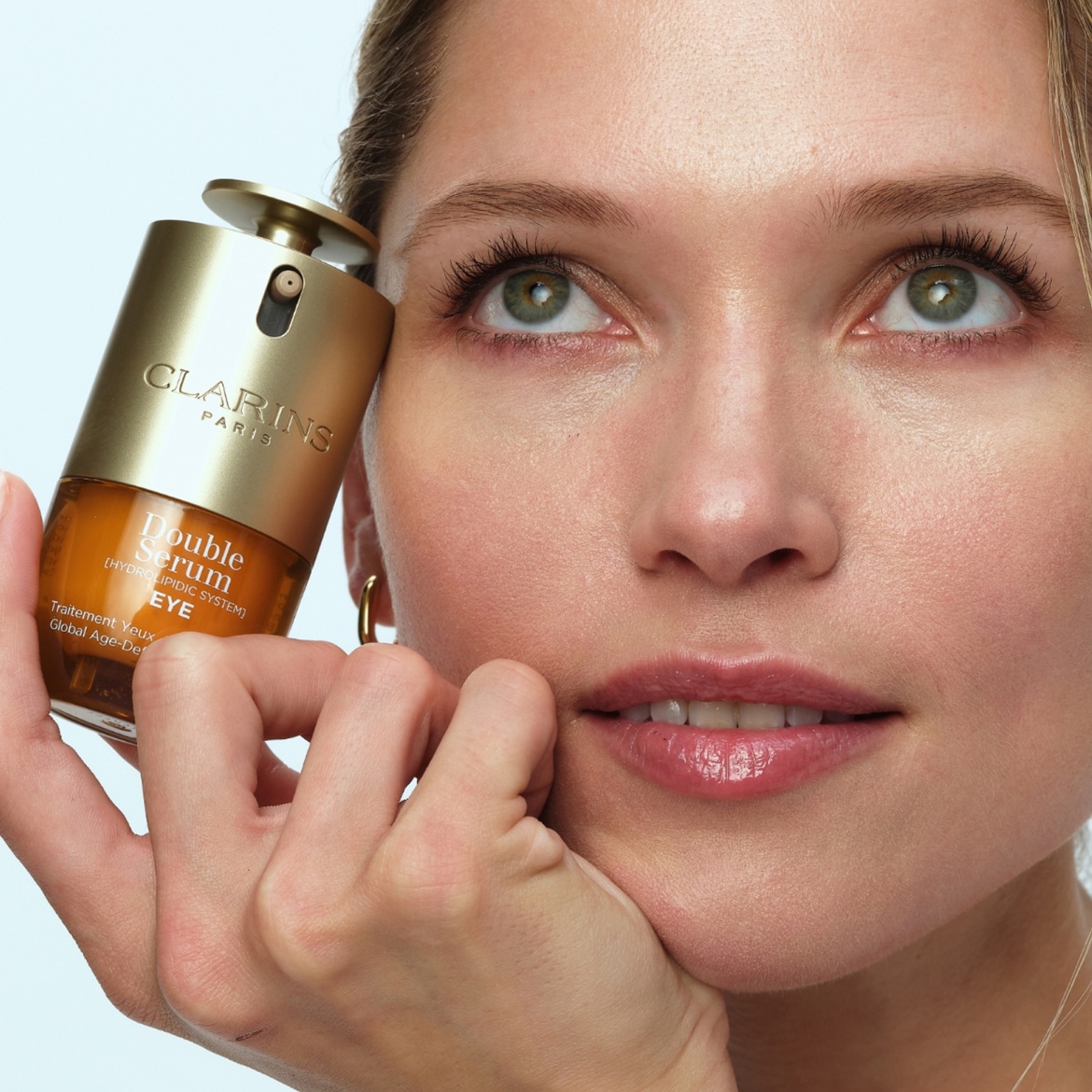 Une femme aux yeux verts tient un flacon de sérum double Clarins doré et ambré près de son visage, le regard tourné vers le haut.
