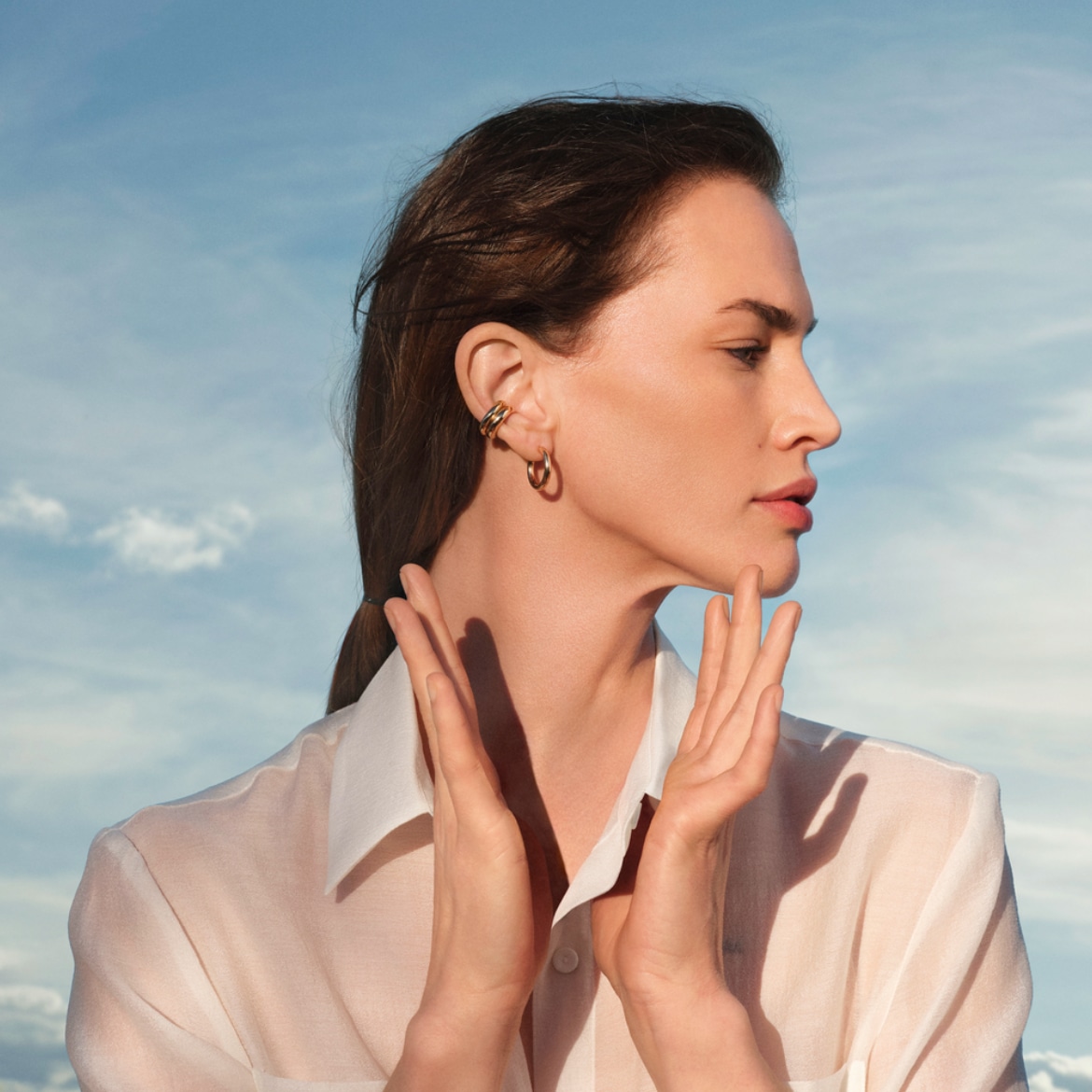 Une femme aux cheveux bruns, vêtue d'une chemise blanche transparente, porte des boucles d'oreilles dorées et des manchettes d'oreille, avec ses mains levées près de son visage, sur un fond de ciel bleu nuageux.