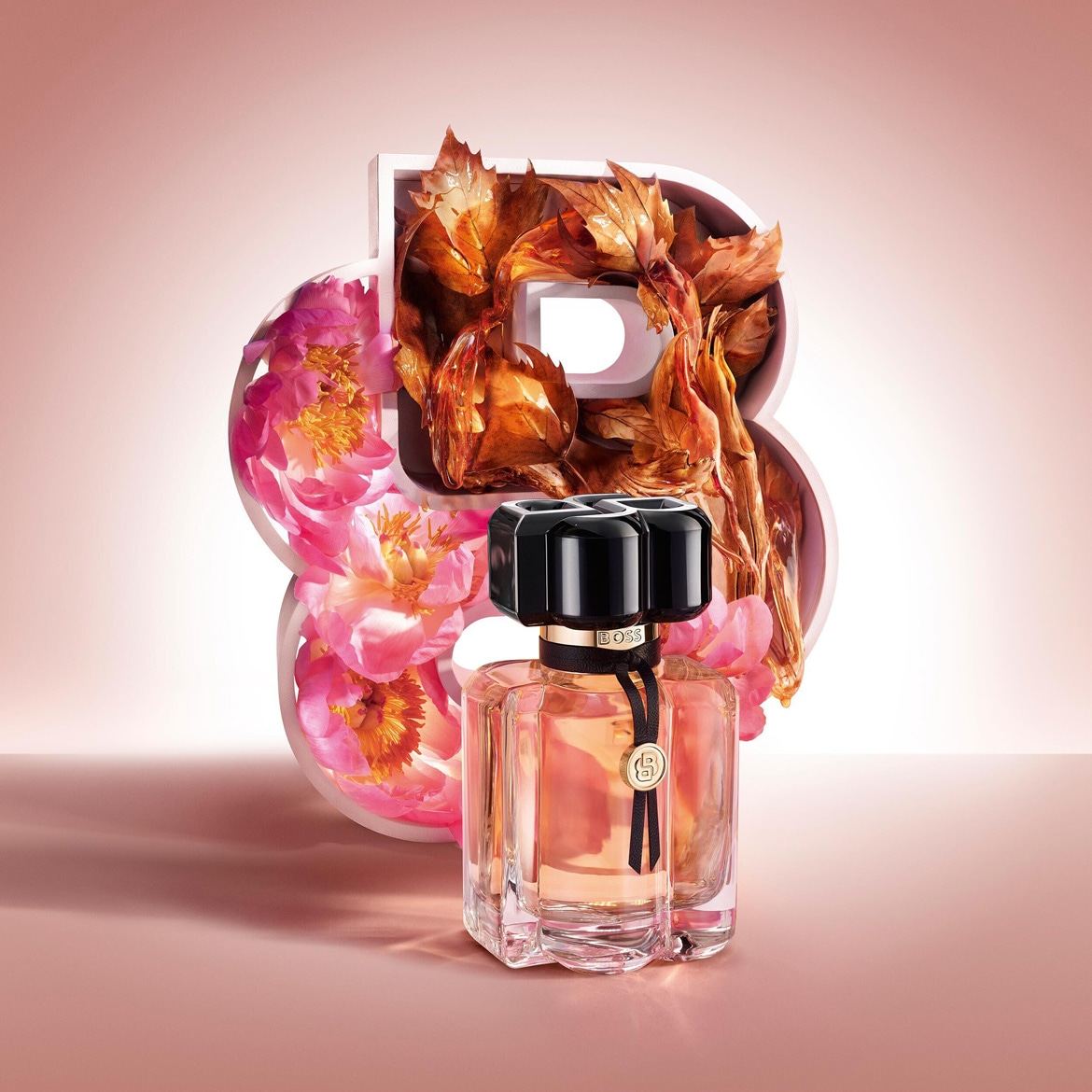 Le parfum Boss The Scent For Her est présenté dans un flacon en verre transparent avec un liquide ambré, un bouchon noir et un ruban noir orné d'un médaillon doré, posé devant un grand chiffre 8 décoré de pivoines roses et de feuilles d'automne.