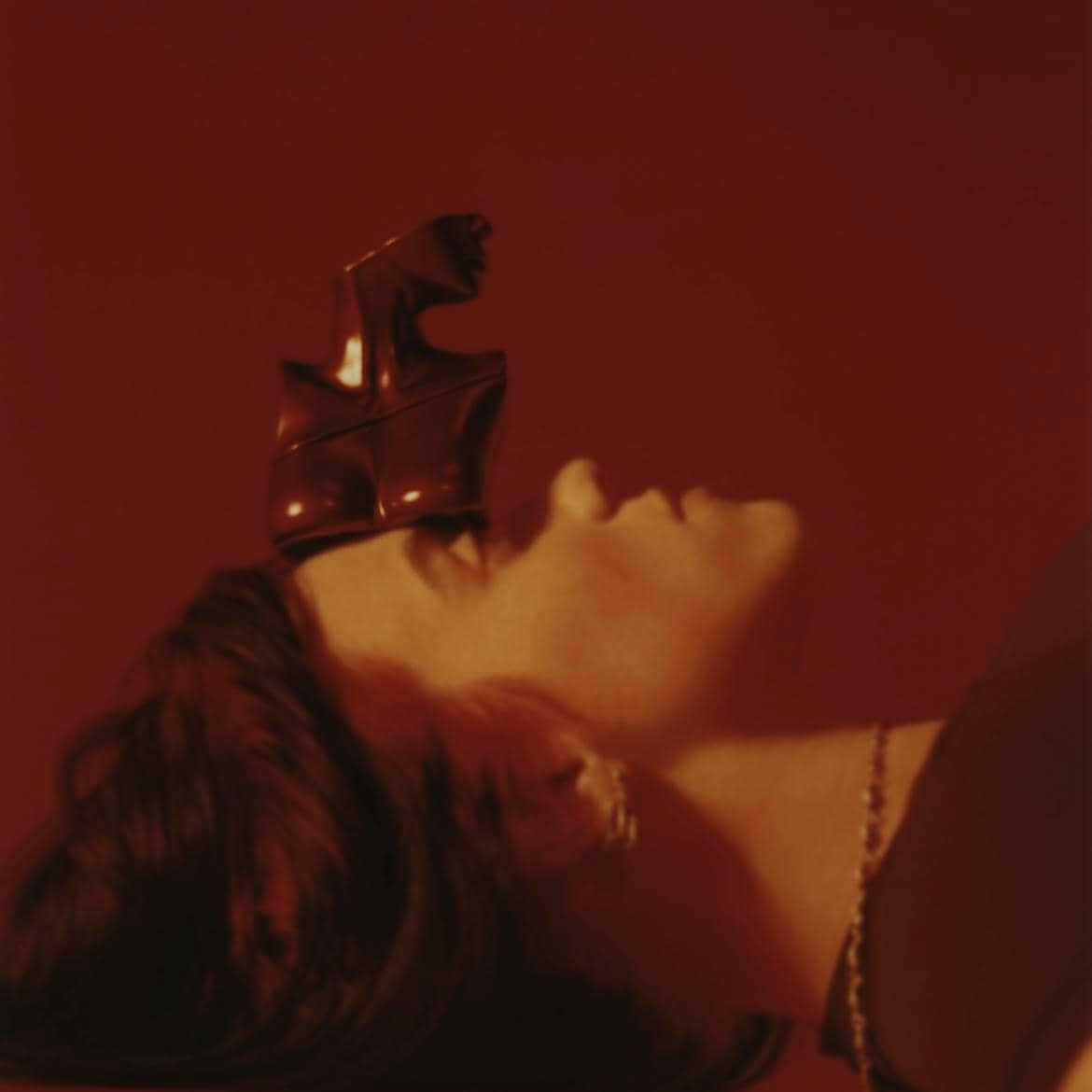 Dans un éclairage rouge profond, une femme est allongée sur le dos, les yeux fermés, avec une sculpture métallique abstraite sur le front.