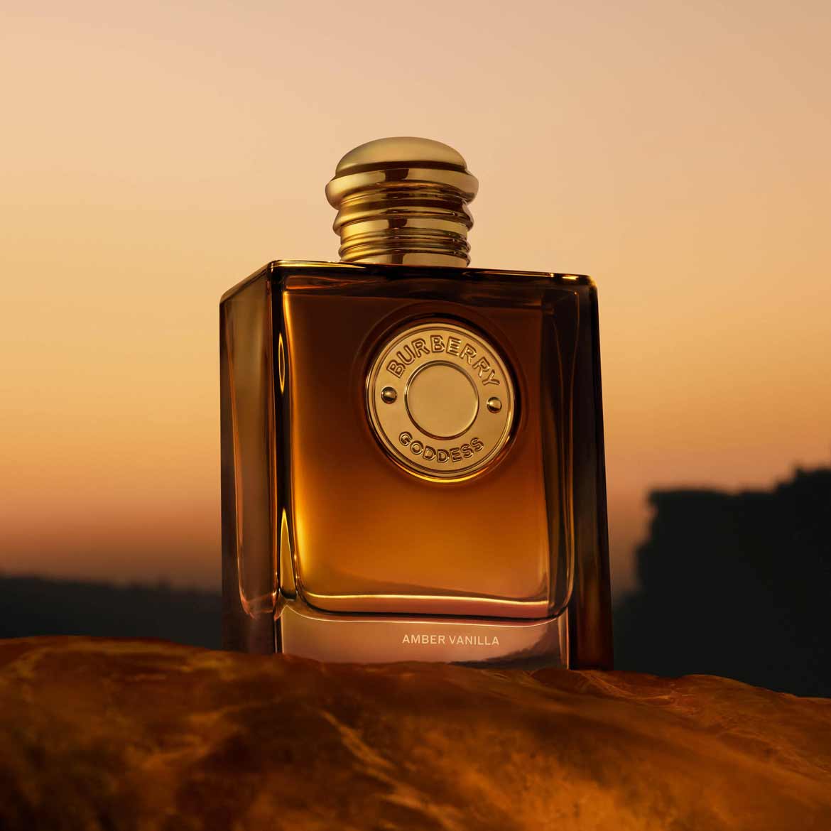 Le parfum Burberry Goddess Ambre Vanille est présenté dans un flacon rectangulaire en verre ambré avec un bouchon doré et un médaillon Burberry Goddess, posé sur une surface texturée aux teintes chaudes.