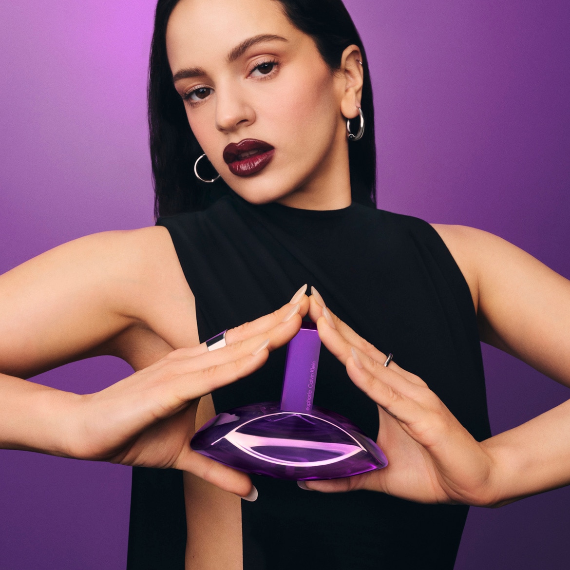 Une femme aux cheveux noirs et aux lèvres bordeaux tient un flacon de parfum violet Euphoria de Calvin Klein, sur un fond violet.