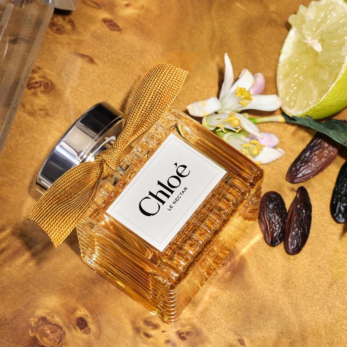 Le parfum Chloé Le Nectar, dans un flacon en verre doré avec un ruban texturé, est présenté avec des fleurs blanches, une tranche de citron vert et des dattes séchées sur une surface en bois.
