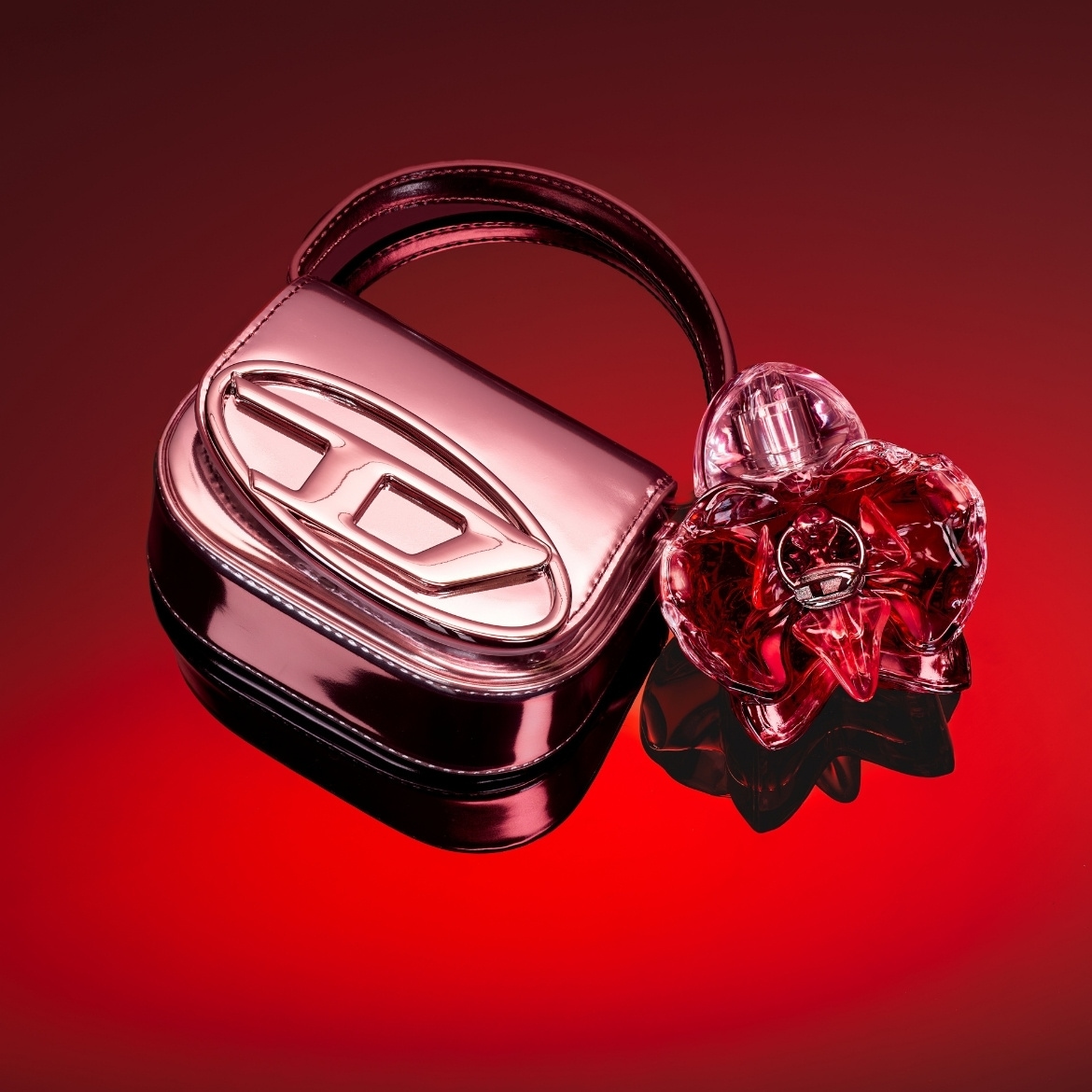 Un sac à main rose métallisé avec le logo Diesel et un flacon de parfum rouge en forme de cœur sont posés sur une surface réfléchissante rouge.