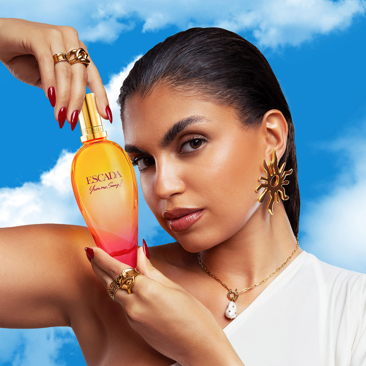 Découvrez le parfum Escada Yumme Sunny dans son flacon dégradé jaune-orange-rose, présenté par un modèle portant des bijoux dorés sur un fond de ciel bleu nuageux.