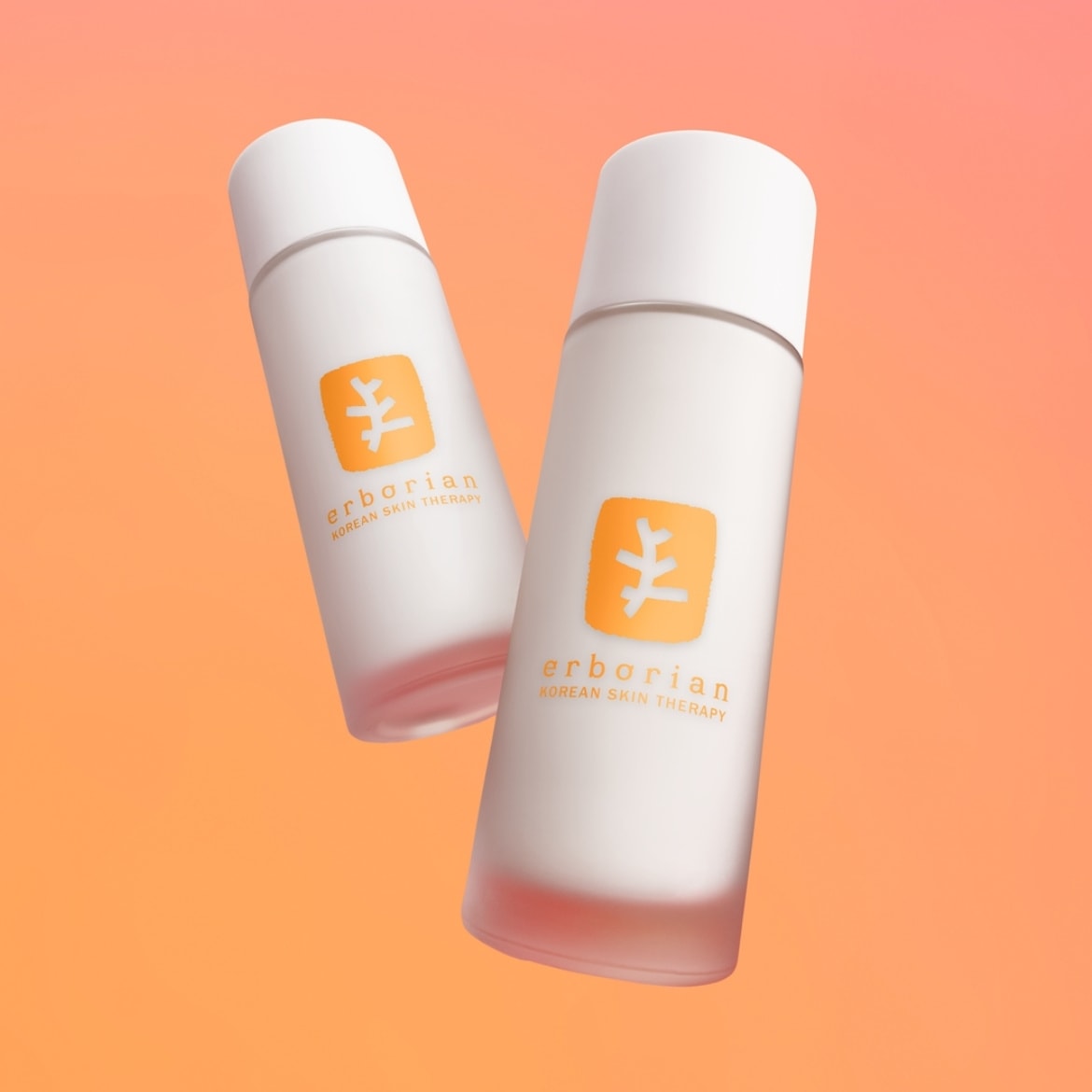 Deux flacons blancs Erborian Korean Skin Therapy avec des bouchons blancs et des étiquettes orange vif, flottant sur un fond dégradé d'orange clair à foncé.