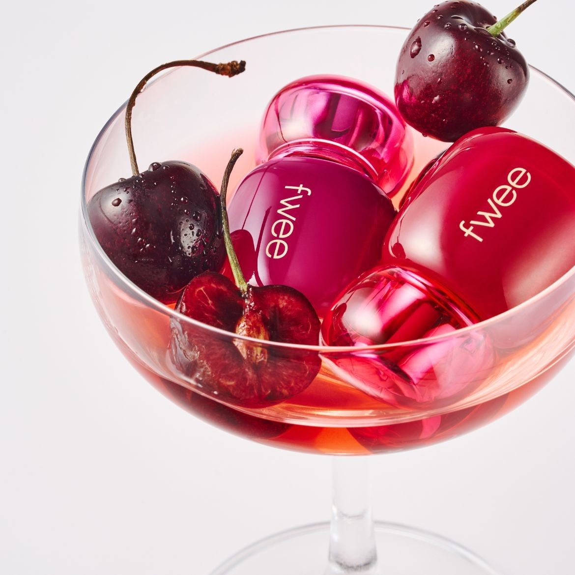 Trois flacons de parfum Fwee de couleur cerise sont présentés dans un verre à cocktail rempli de liquide rose, accompagnés de cerises fraîches et juteuses.