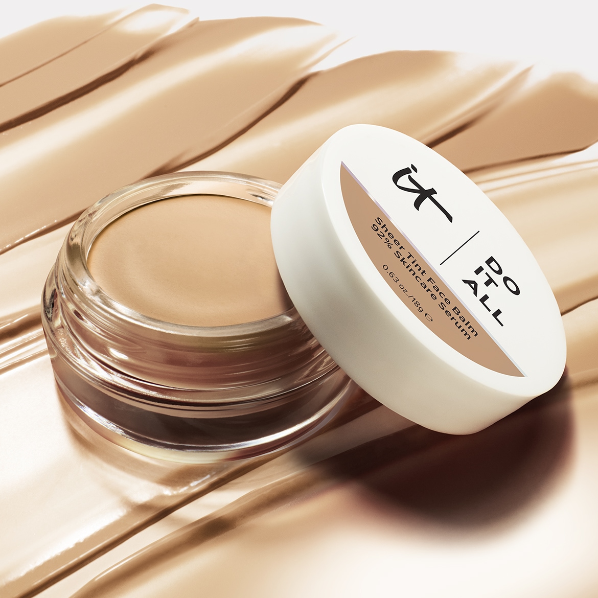 Un pot ouvert de baume teinté IT Cosmetics Do It All Sheer Tint Face Balm, 92% Skincare Serum, est posé sur des traces de fond de teint beige clair, révélant sa texture crémeuse et sa teinte naturelle.