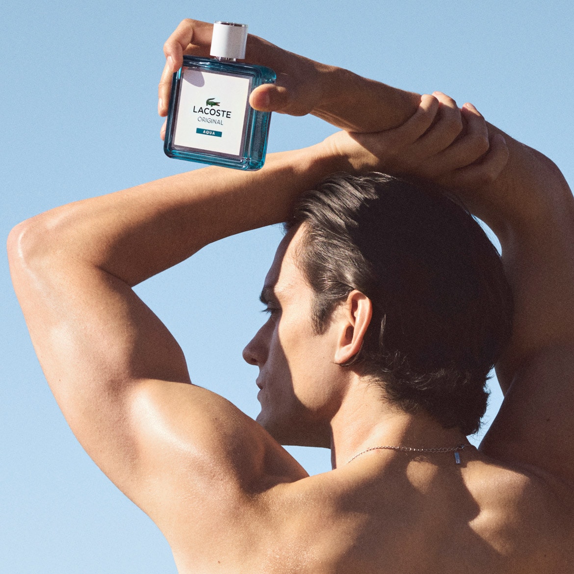 Un homme torse nu tient au-dessus de sa tête un flacon de parfum Lacoste Original Aqua, un parfum frais et aquatique pour homme, sur un fond de ciel bleu clair.