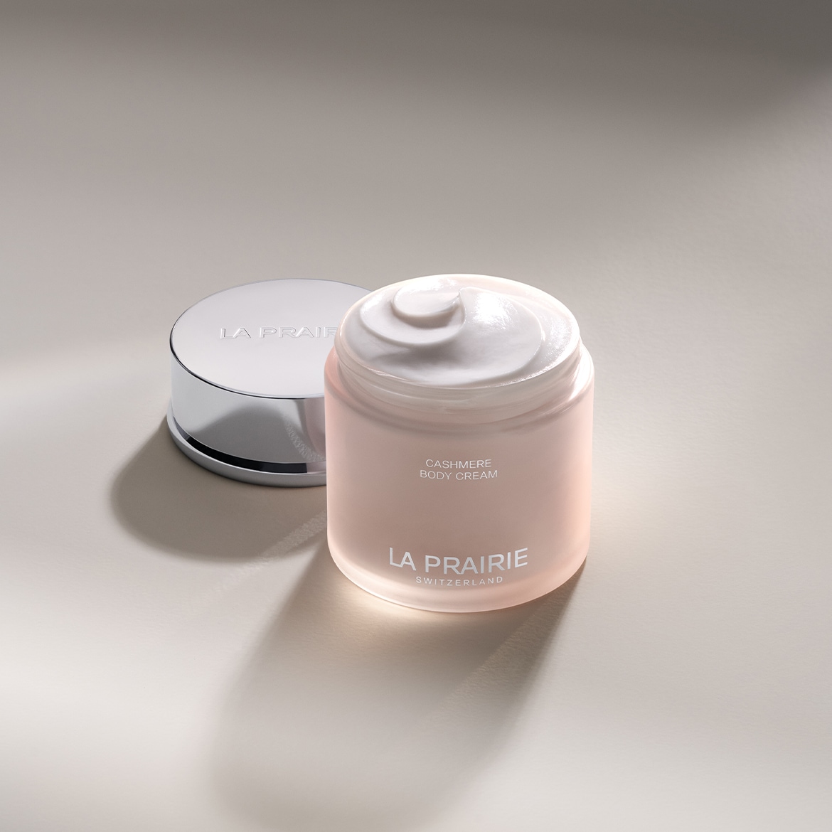 Découvrez la Crème Corps Cachemire de La Prairie, présentée dans un pot rose doux avec un couvercle argenté réfléchissant, offrant une hydratation luxueuse pour une peau douce et soyeuse.