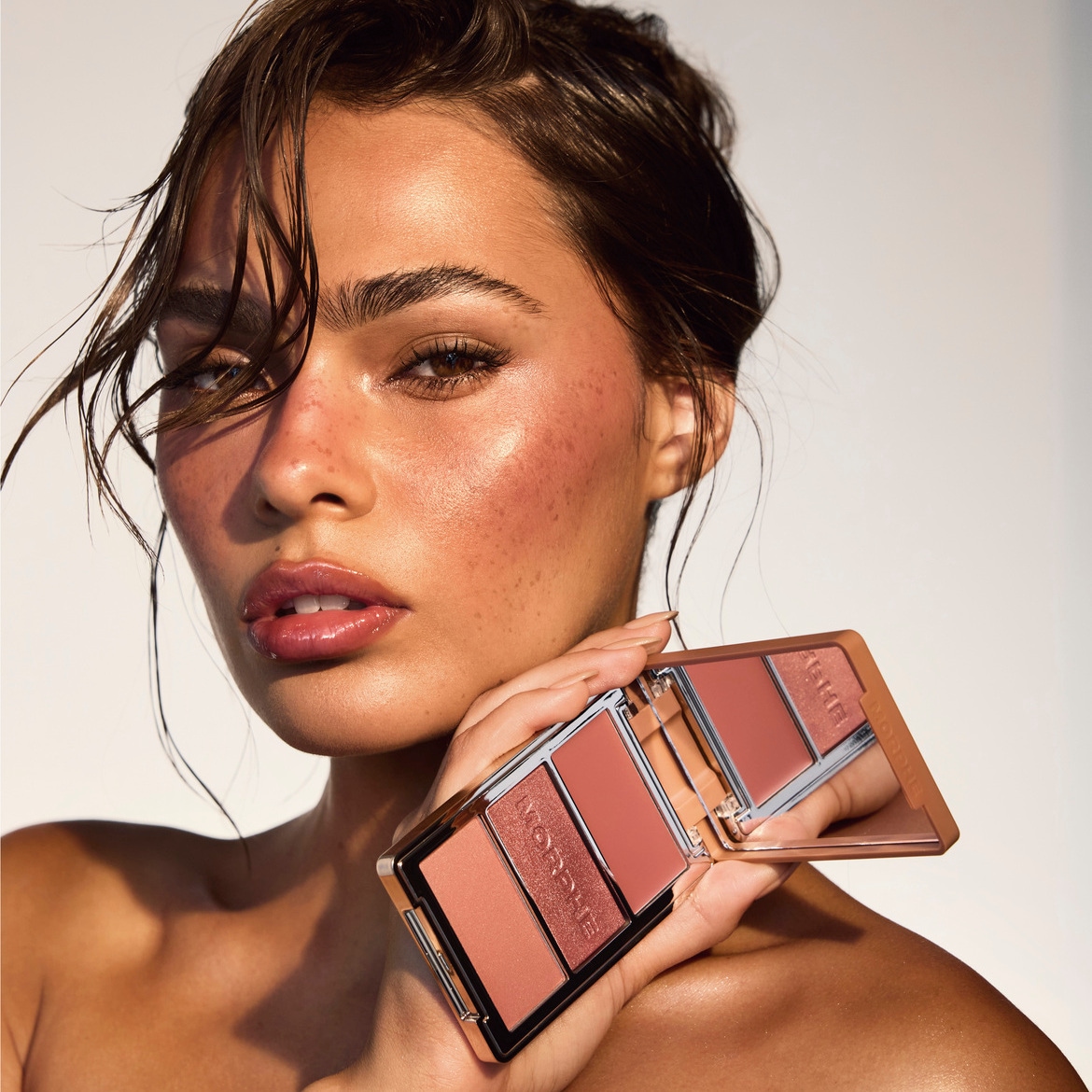 Une femme au teint hâlé et aux cheveux mouillés tient une palette de maquillage Morphe avec trois teintes de blush et d'highlighter, reflétant un look frais et lumineux.