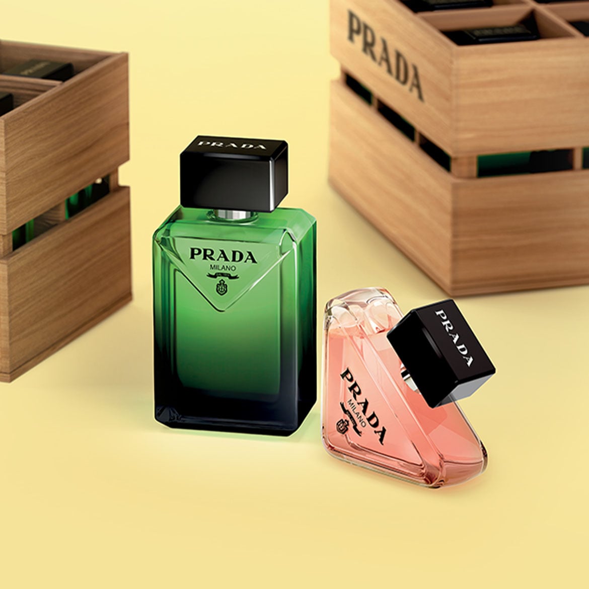 Découvrez les parfums Prada dans leurs flacons distinctifs : l'un vert et l'autre rose, coiffés de bouchons noirs élégants, présentés à côté de caisses en bois.