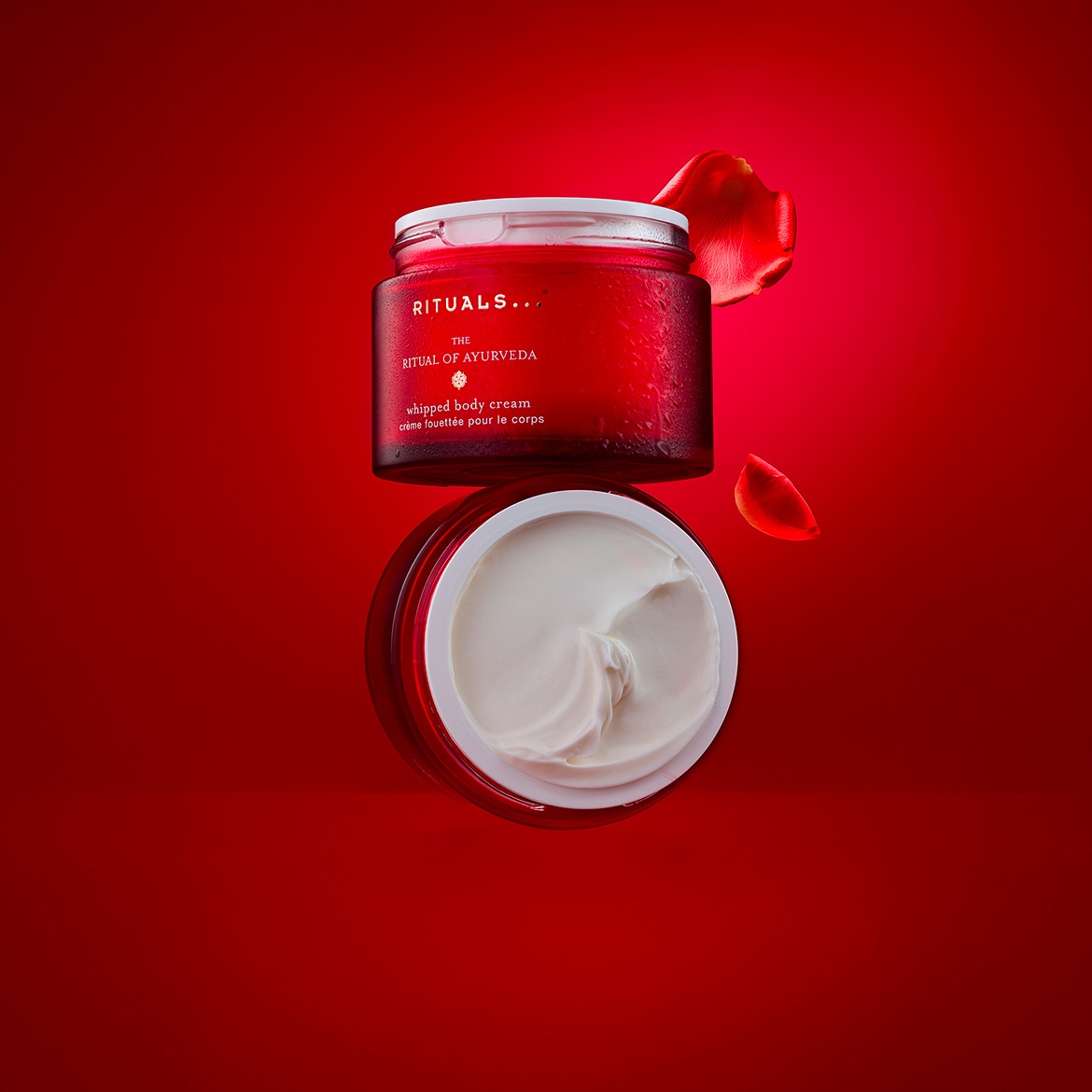 Découvrez le Rituel de l'Ayurveda de Rituals... avec cette crème fouettée pour le corps présentée dans un pot rouge et blanc, accompagnée de pétales de rose rouges.