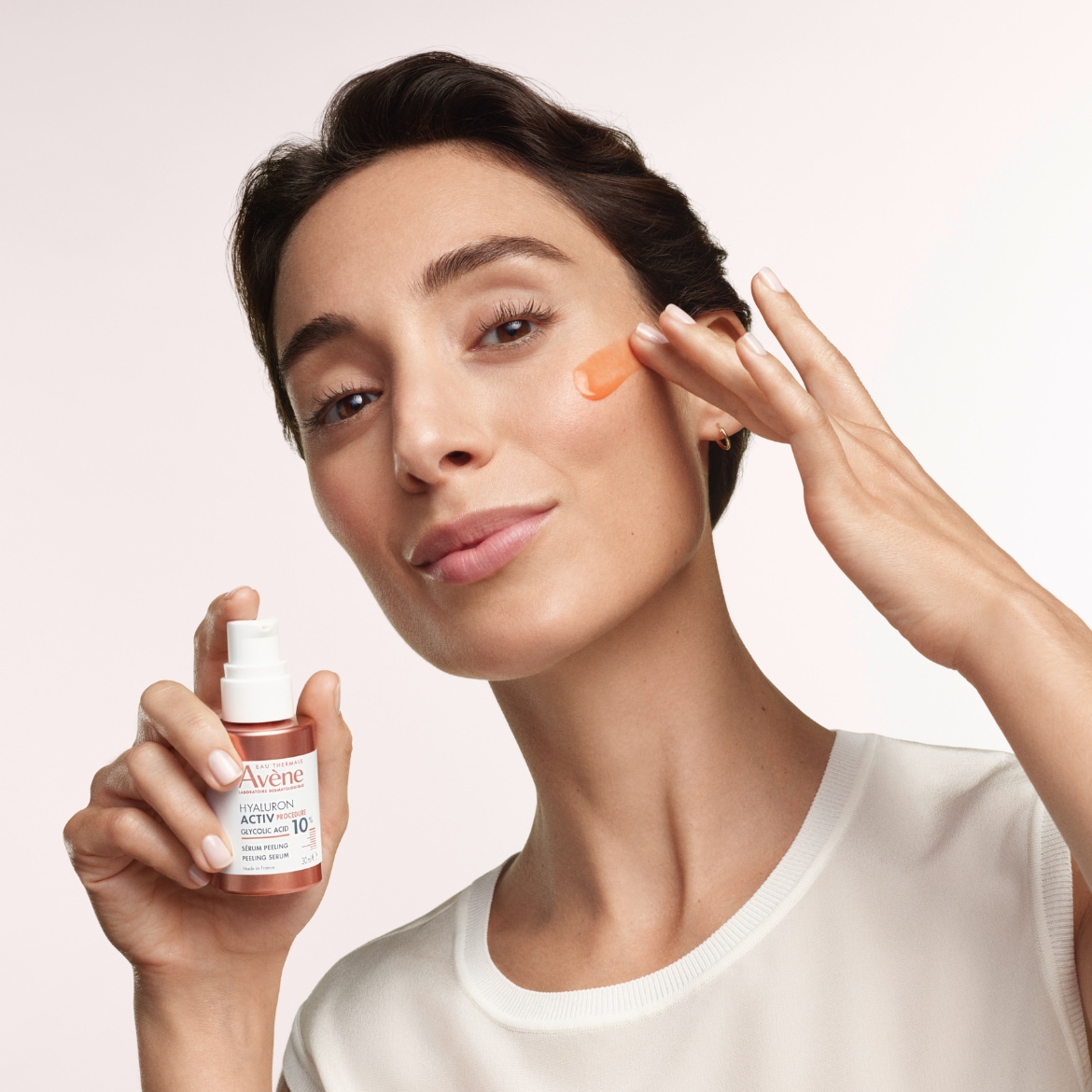 Une femme applique le sérum peeling Avène Hyaluron Activ B3 sur sa joue, le flacon rose saumon à la main, pour une peau éclatante.