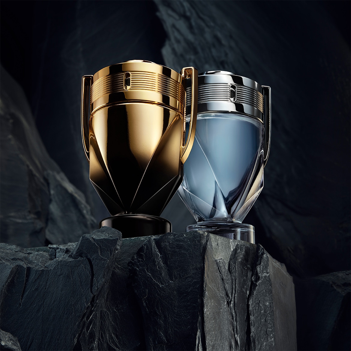 Deux flacons de parfum Invictus en forme de trophée, l'un doré et l'autre argenté, posés sur un rocher sombre, évoquant la victoire et le luxe.