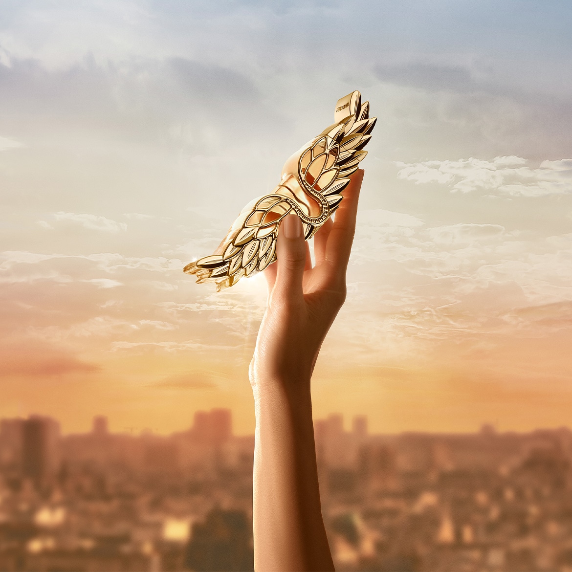 Main tend une sculpture dorée en forme d'aile sur fond de ciel nuageux au coucher du soleil, symbolisant l'élégance et la liberté.
