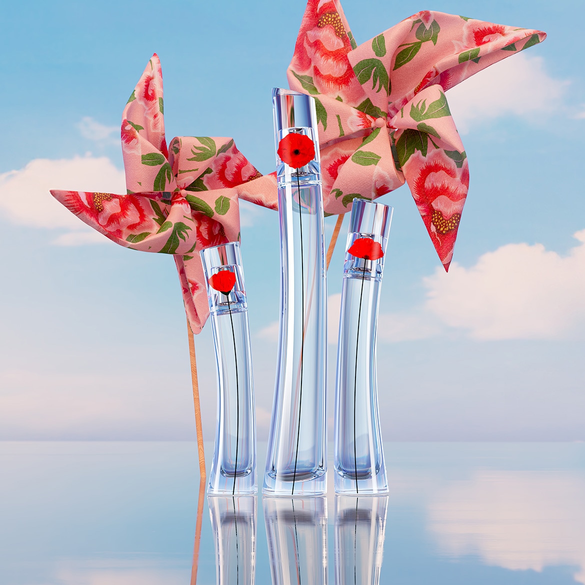 Trois flacons de parfum transparents avec un bouchon rouge en forme de coquelicot, accompagnés de deux moulins à vent roses à motifs floraux, évoquant un parfum frais et printanier.