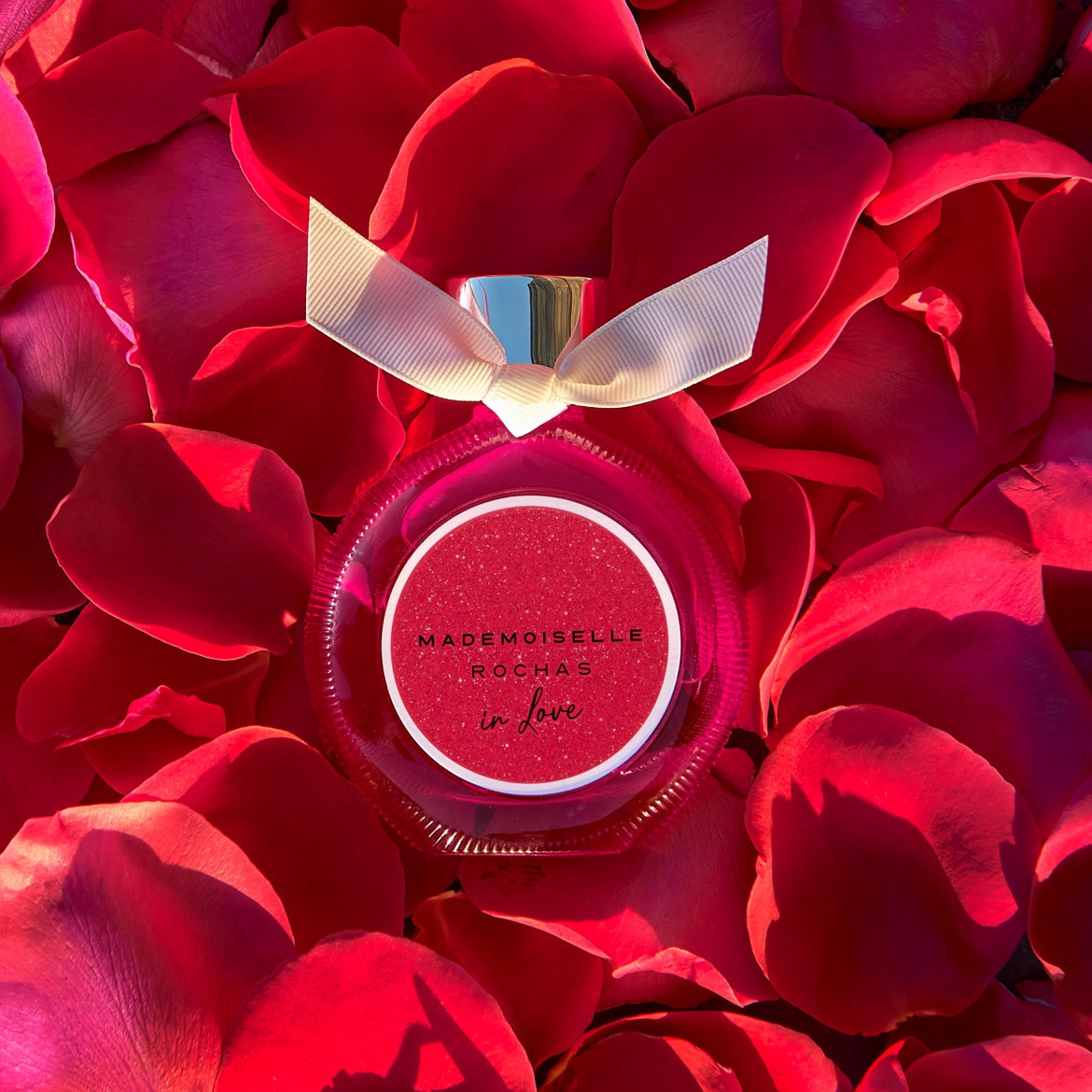 Découvrez Mademoiselle Rochas In Love, un parfum floral fruité pétillant, présenté dans un flacon rose délicat orné d'un ruban ivoire, sur un lit de pétales de rose rouge.