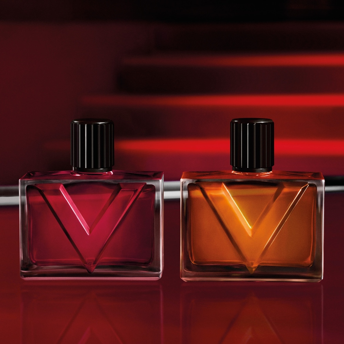 Deux flacons de parfum rectangulaires avec des capuchons noirs cannelés, l'un rempli d'un liquide rouge et l'autre d'un liquide ambré, sont posés sur une surface réfléchissante rouge, avec un arrière-plan rouge flou.