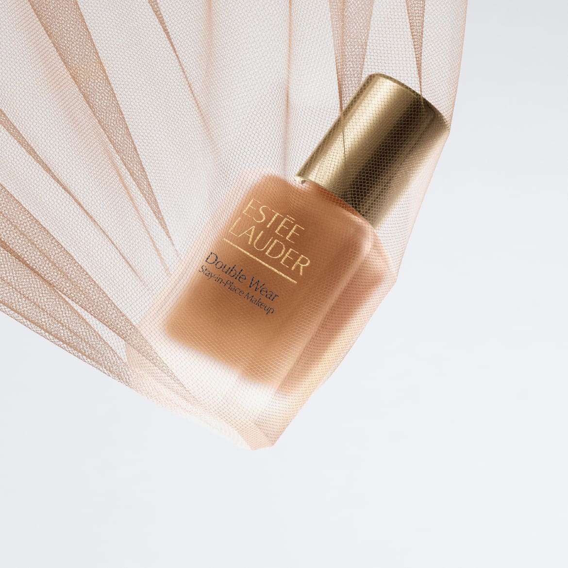 Flacon de fond de teint Estée Lauder Double Wear couleur pêche, coiffé d'un bouchon doré, enveloppé dans un tissu en tulle abricot.