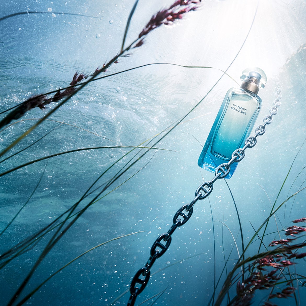 Le parfum Hermès Un Jardin Sous La Mer, un flacon bleu dégradé, est suspendu par une chaîne argentée dans un décor aquatique avec des herbes marines, évoquant une fraîcheur et une évasion sous-marine.