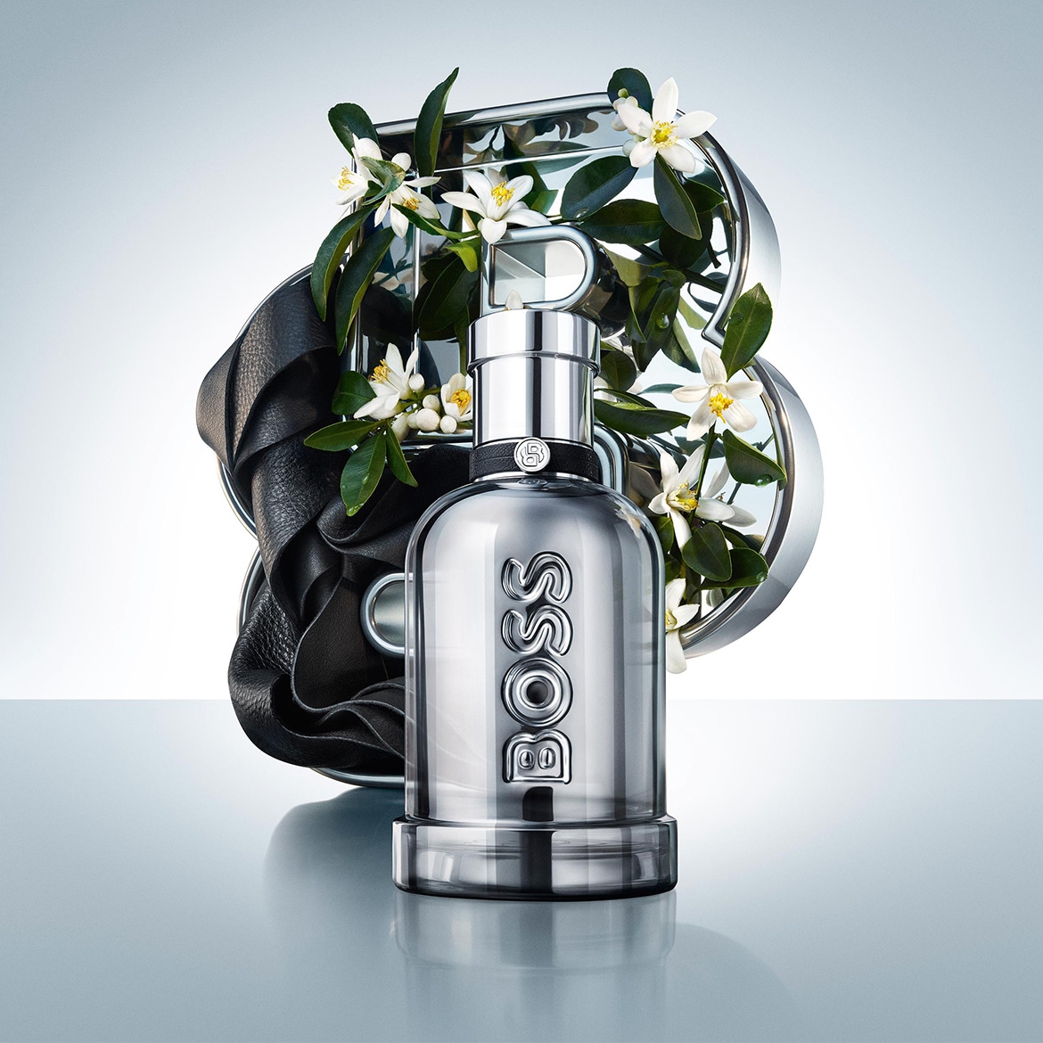 Le parfum BOSS The Scent Le Parfum pour Homme est présenté dans un flacon en verre argenté avec des fleurs blanches et des feuilles vertes, évoquant une fragrance fraîche et élégante.